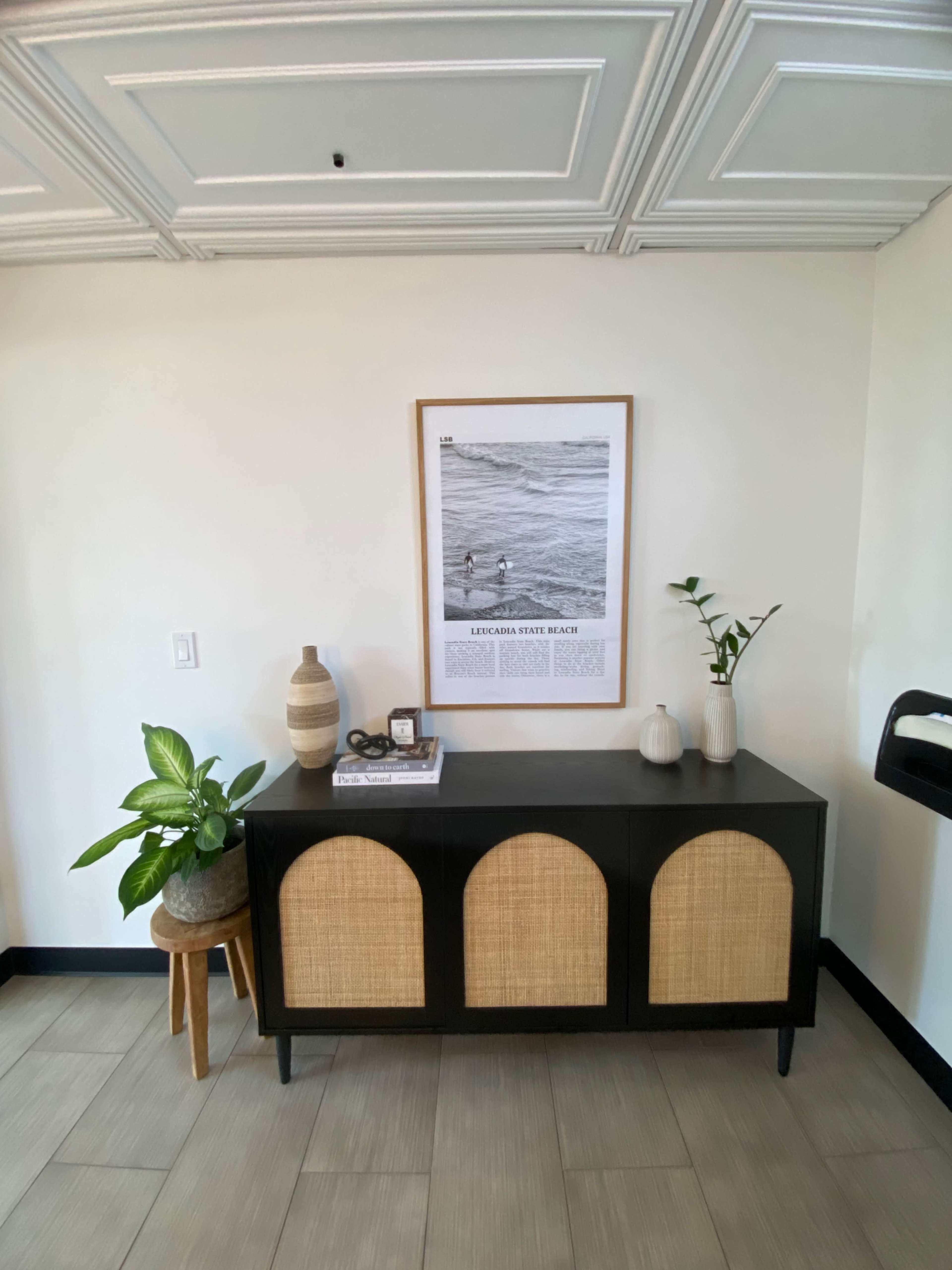 A sideboard with three arched doors sits against a wall, featuring a framed black-and-white print and decorative plants.