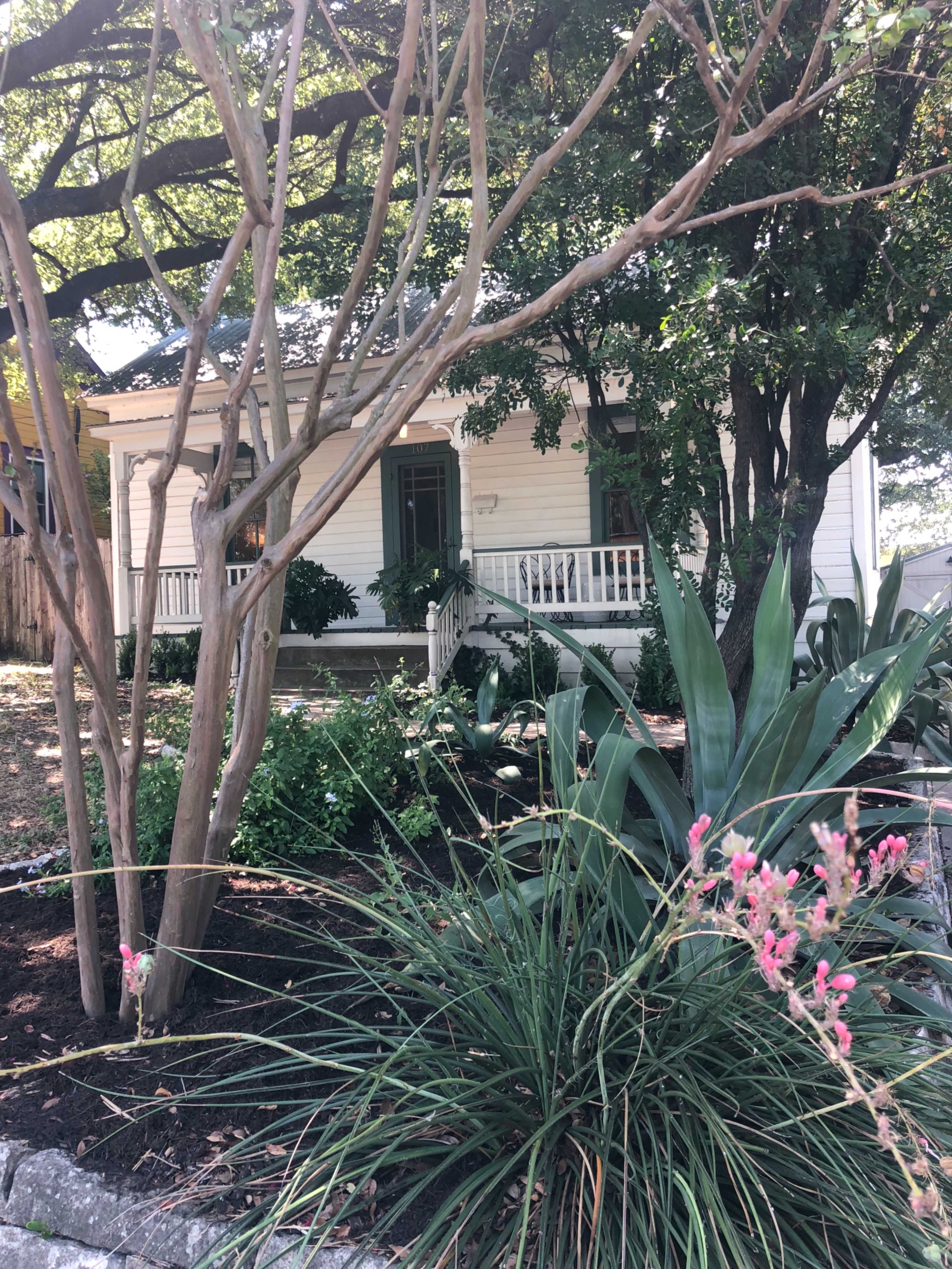 A white house with a porch is set back among trees and shrubs in a garden.