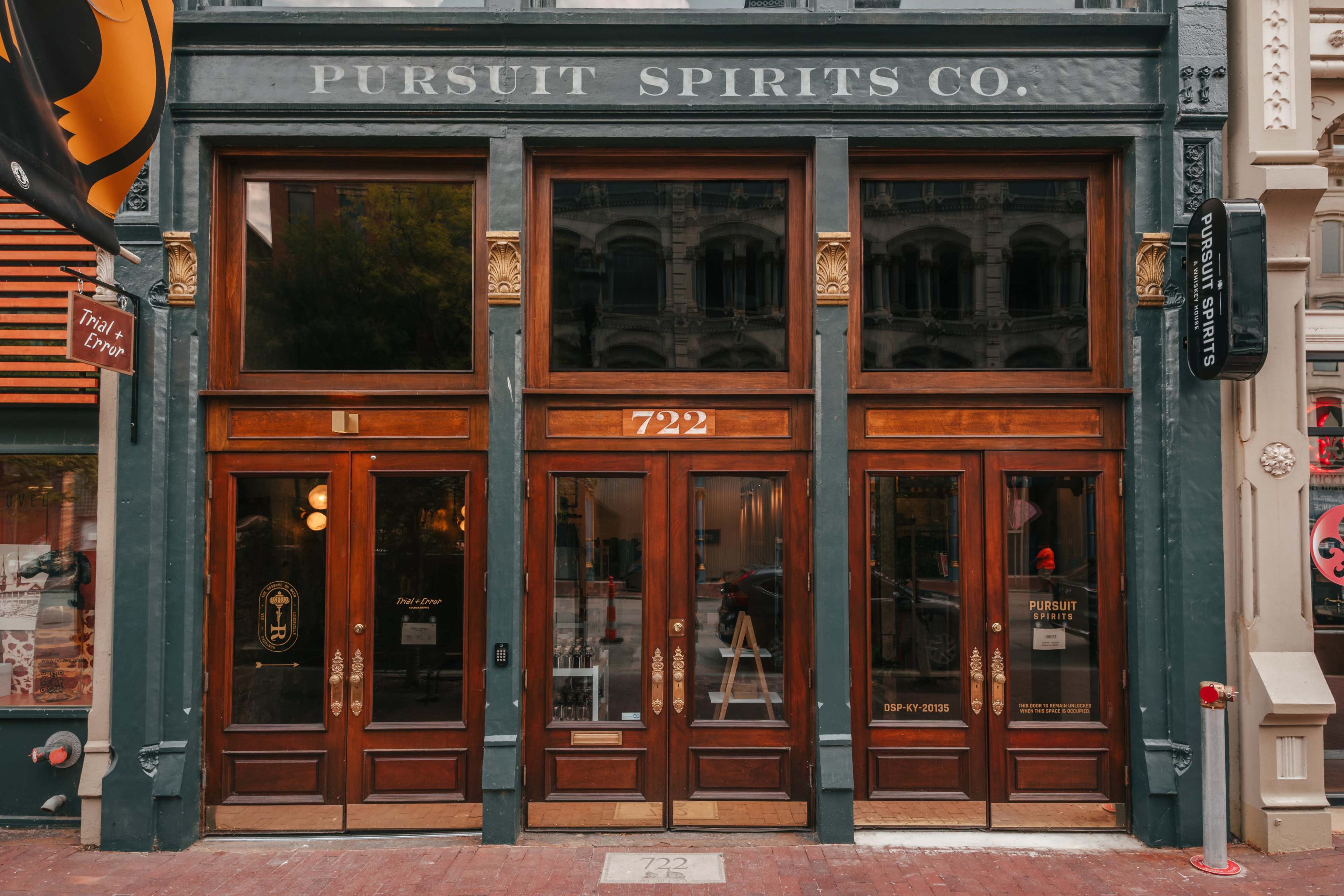 The image shows the exterior of a building featuring large wooden doors with glass panels, labeled "Pursuit Spirits Co." and displaying the address "722."