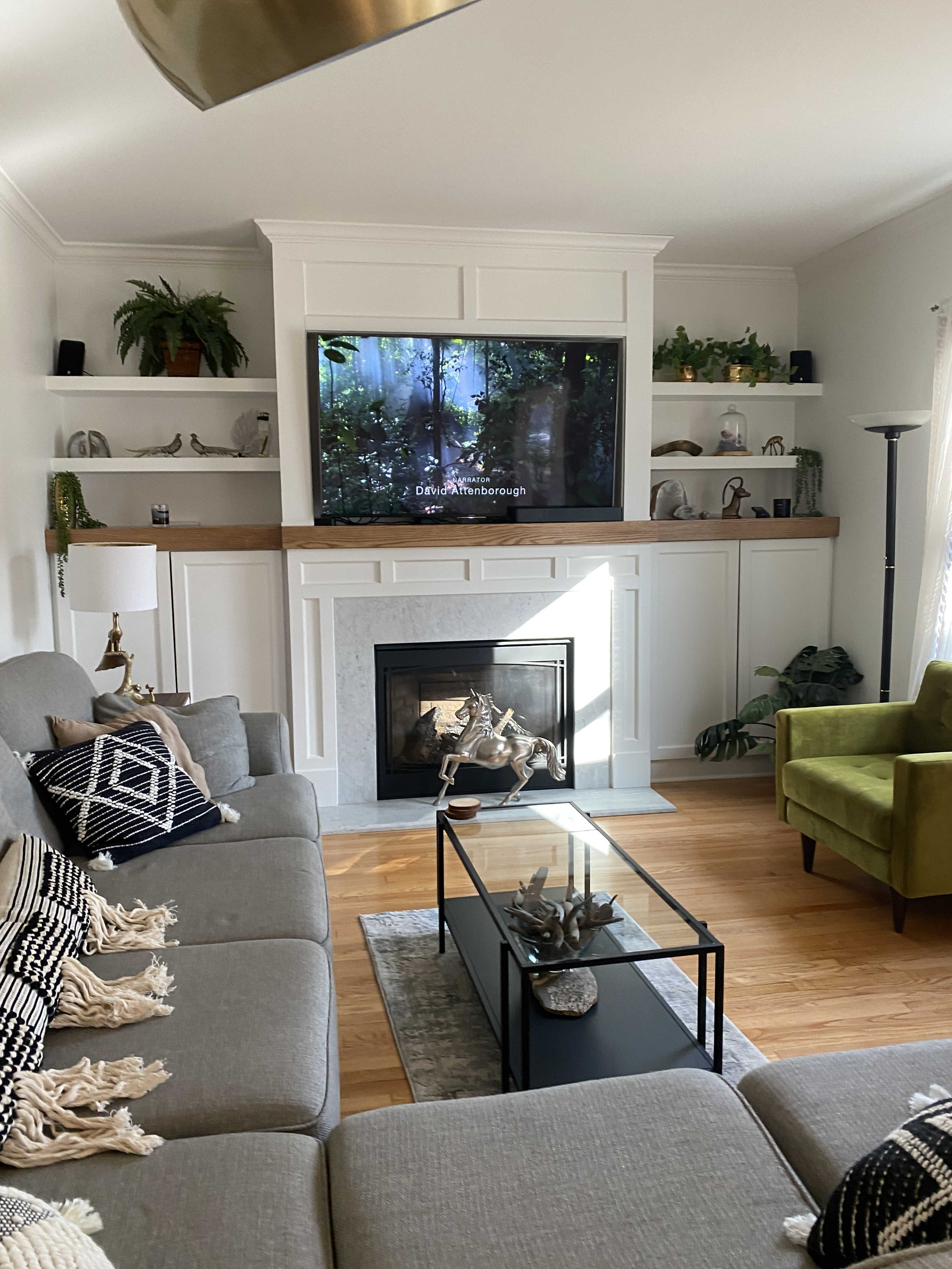 A living room features a gray sectional sofa, a glass coffee table, a green accent chair, and a television mounted above a white fireplace with decorative shelves.