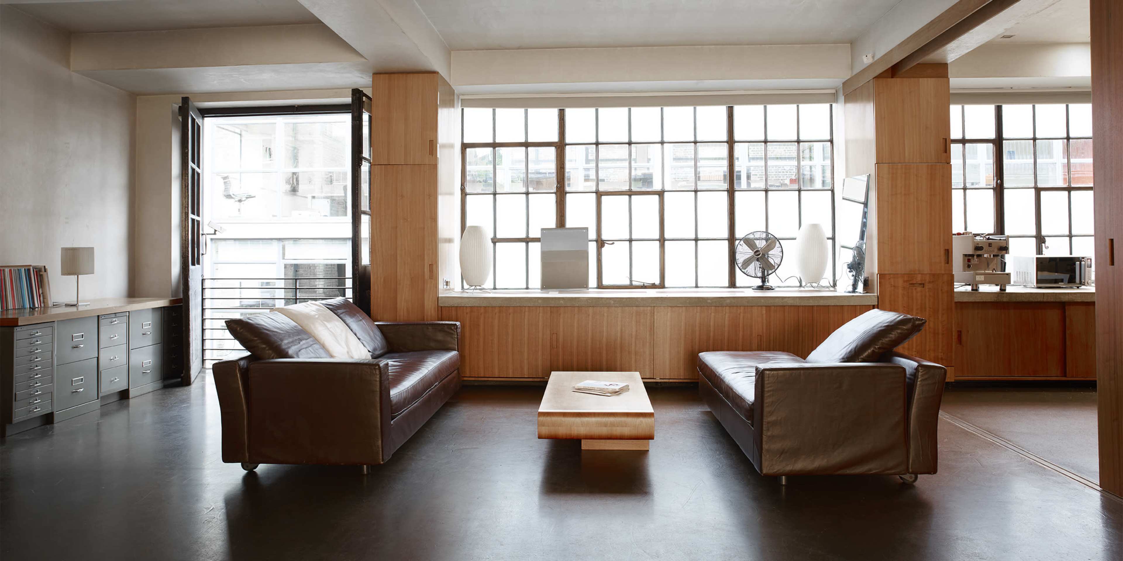 A modern living area features two leather sofas facing a wooden coffee table, with large windows providing natural light.