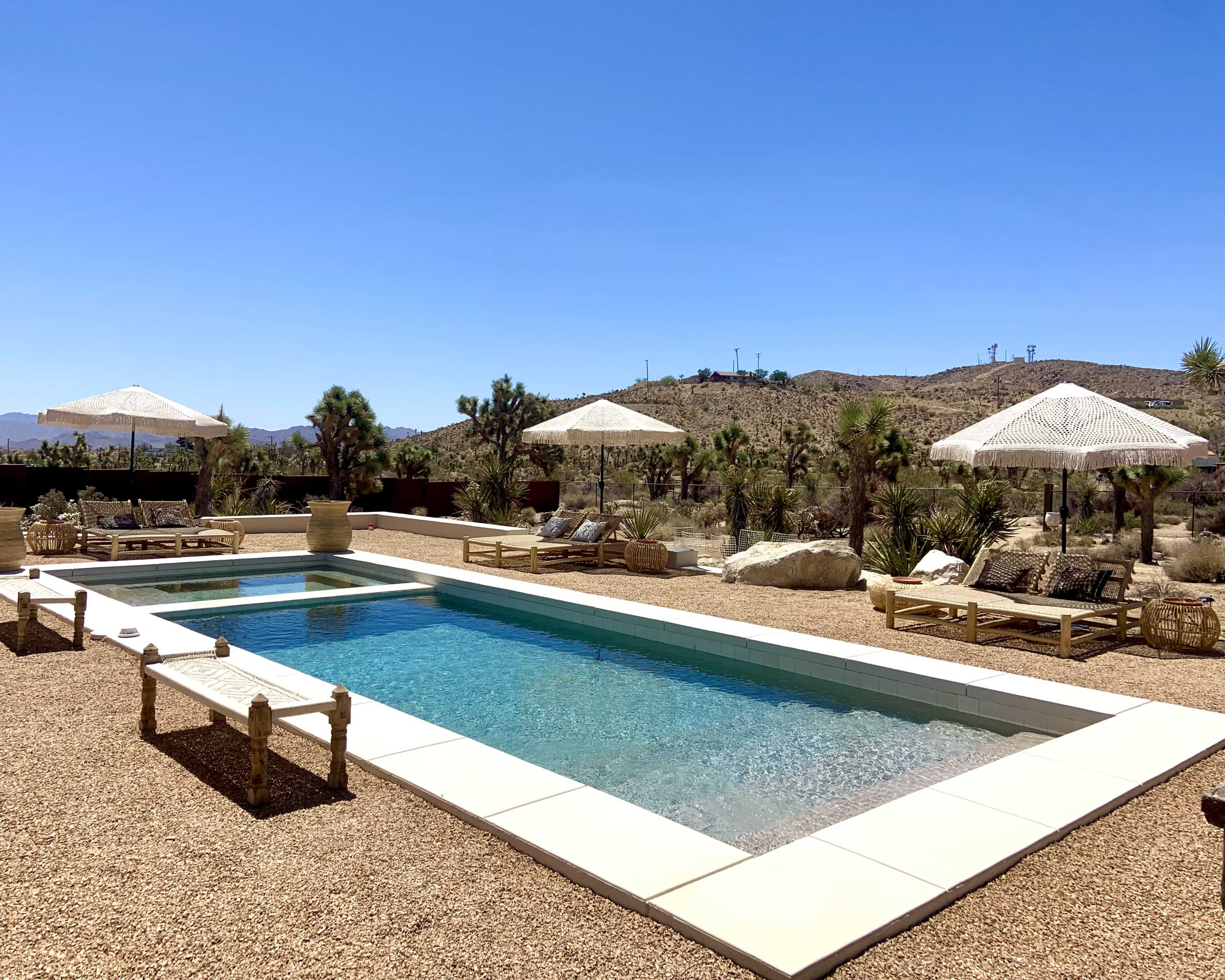 The image shows a desert landscape featuring a swimming pool surrounded by lounge chairs and shaded by parasols.