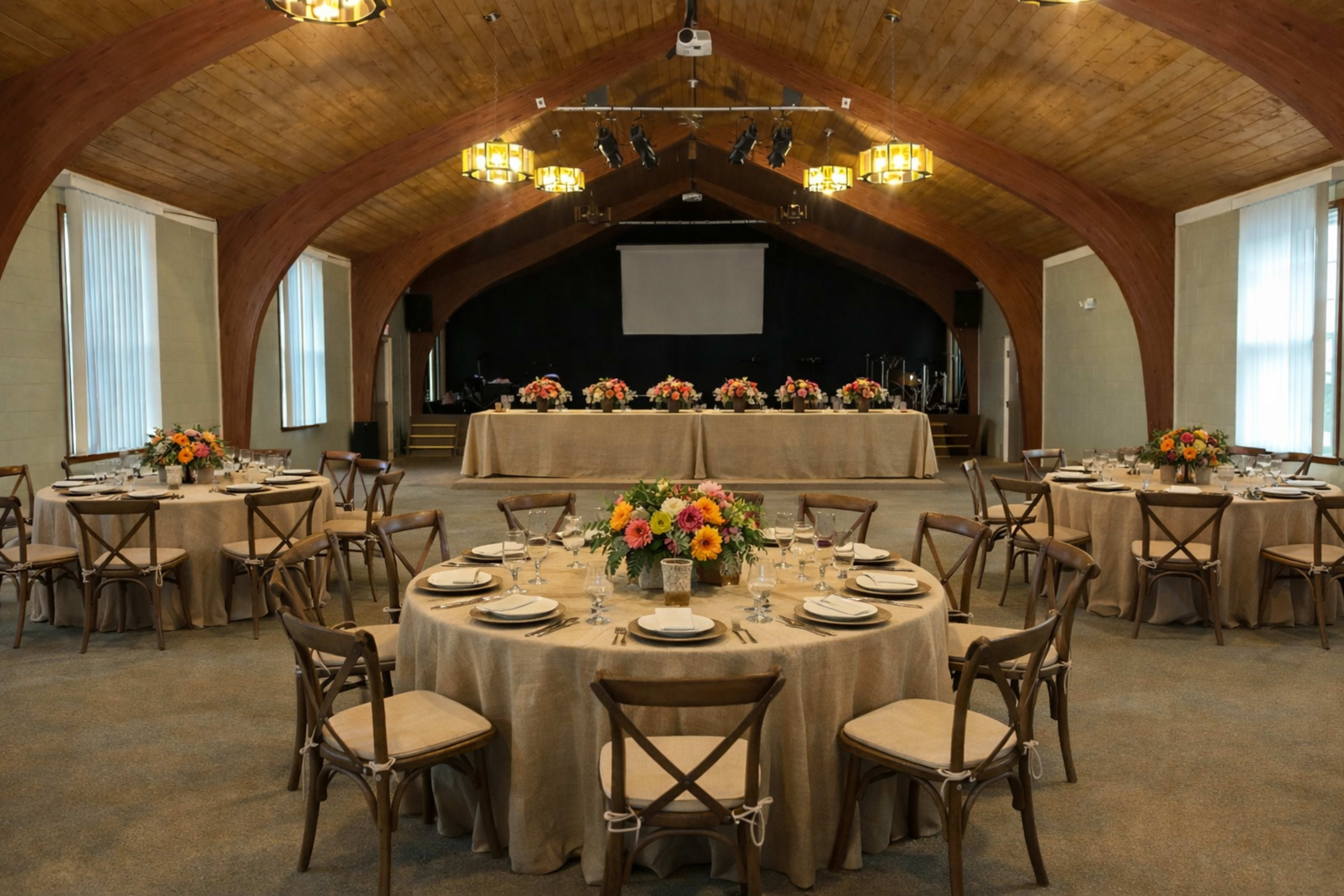 A well-decorated banquet hall features round tables set with floral centerpieces and a stage at the front.