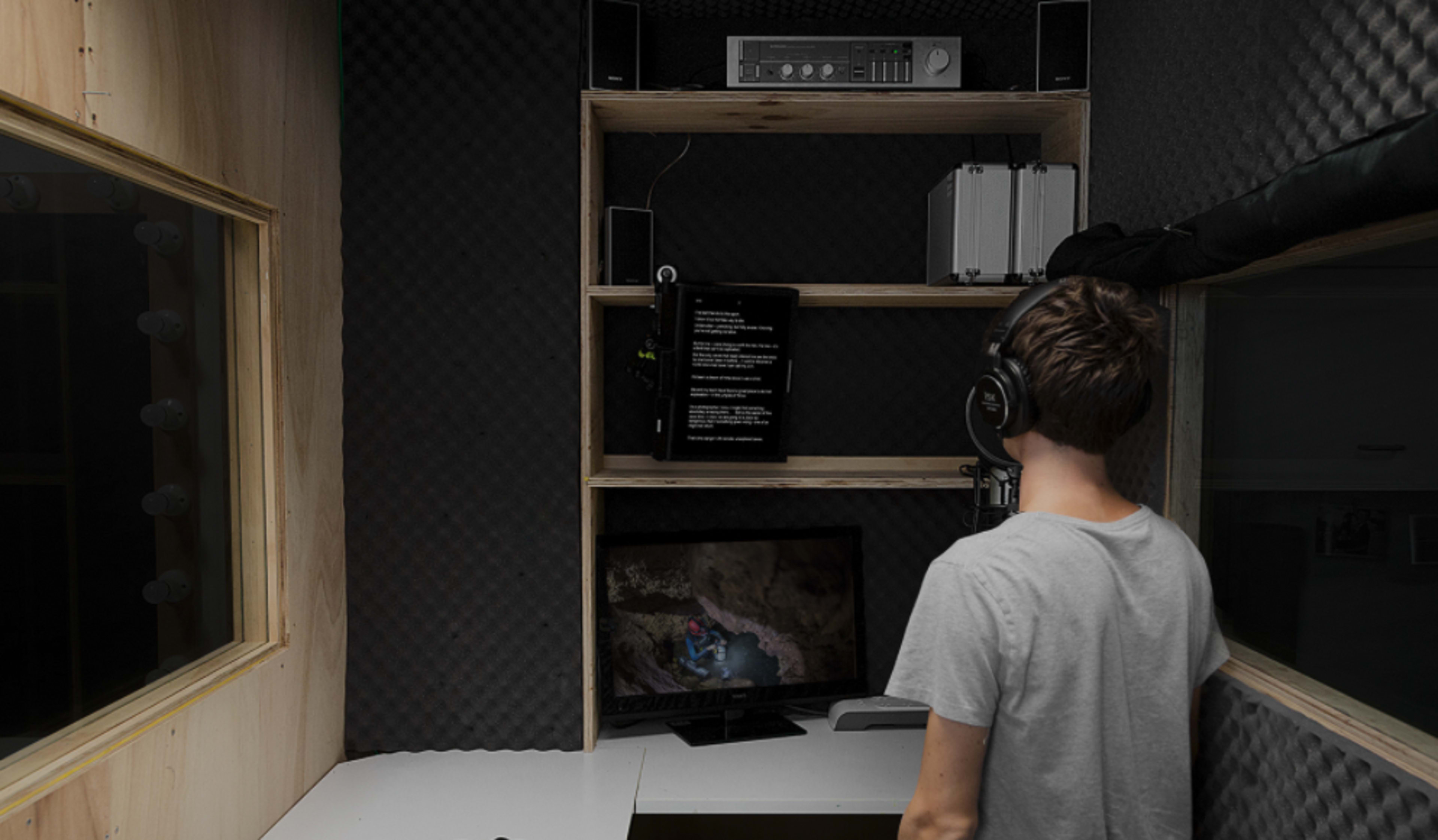 A person wearing headphones stands in a soundproof recording booth, facing a monitor and a shelf with audio equipment.