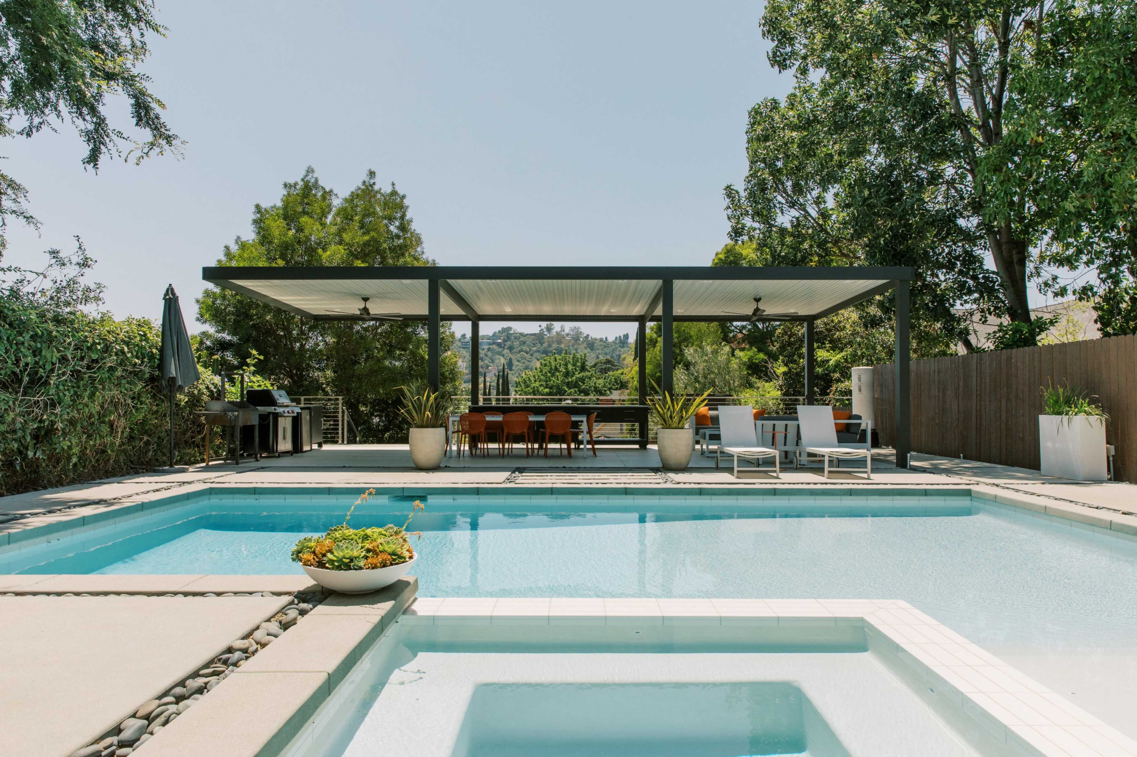 A rectangular swimming pool with a hot tub is surrounded by lounge chairs and a dining area under a covered patio.