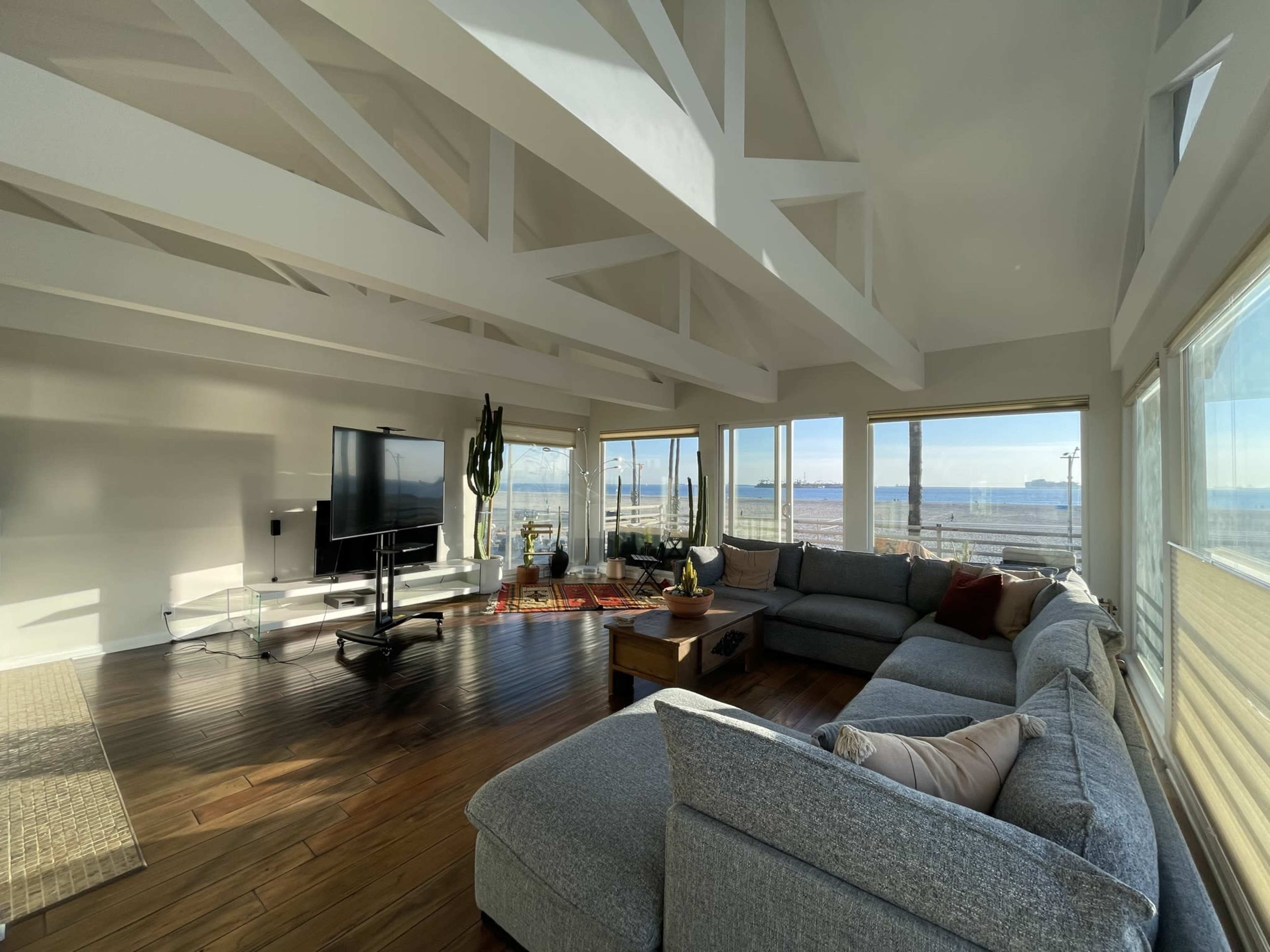 A spacious living room features a large gray sectional sofa, a flat-screen TV, and expansive windows with a view of the water.