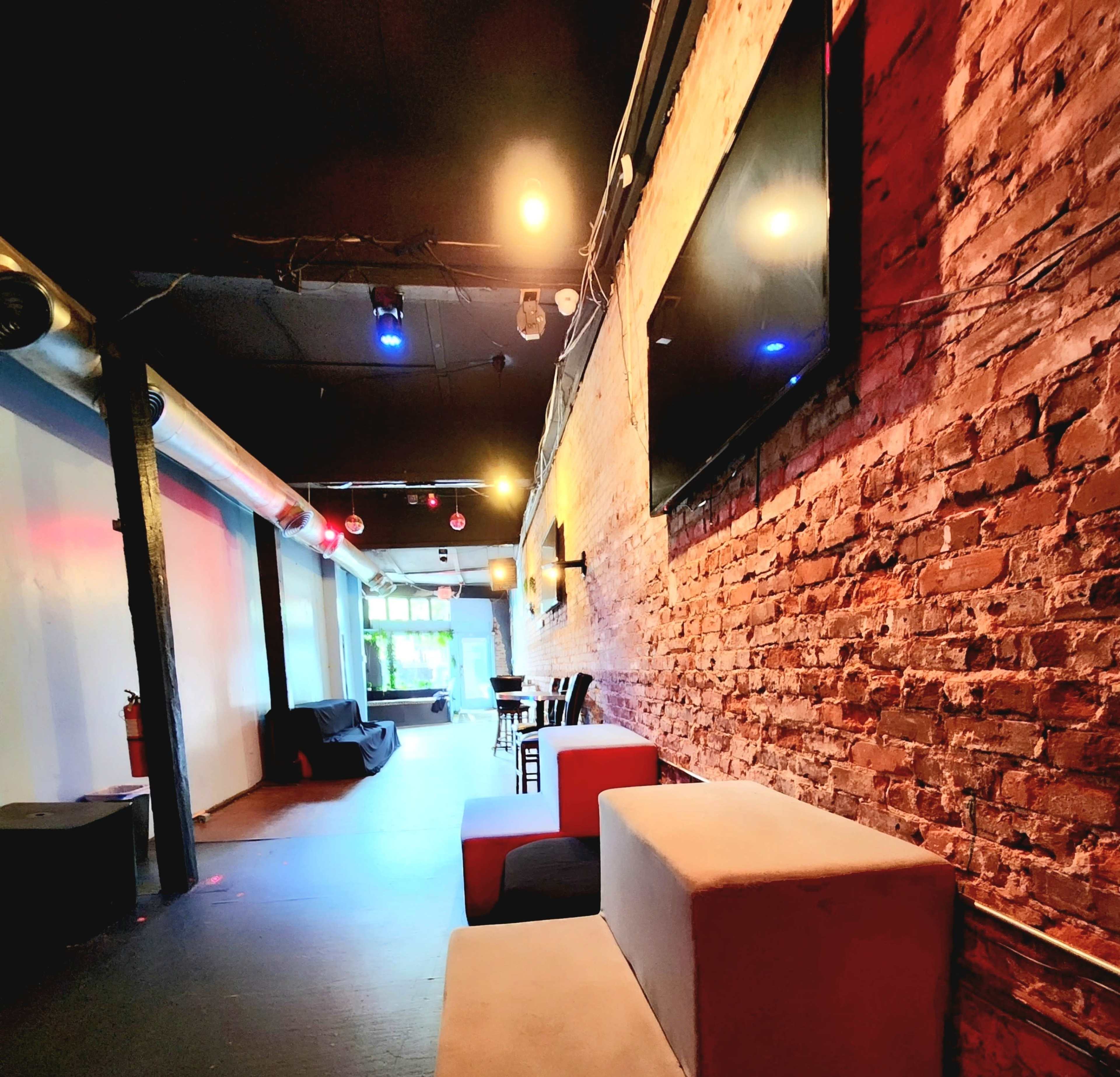 The image shows a narrow indoor space with exposed brick walls, a few seating areas, tables, and soft lighting.