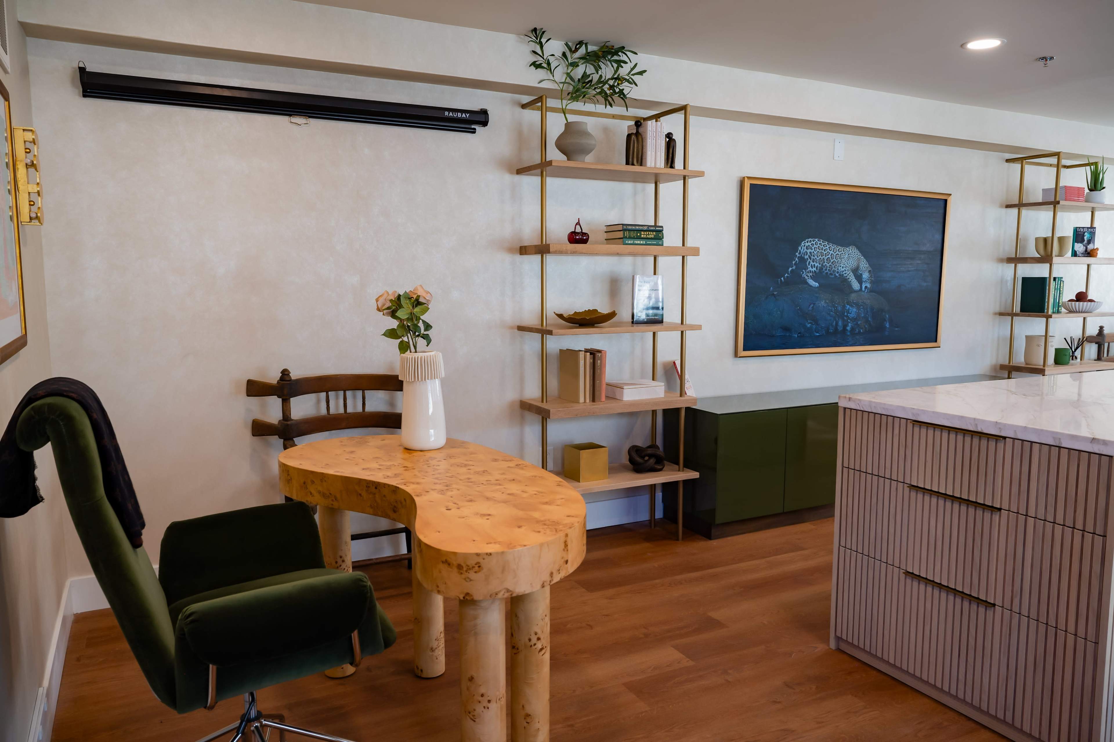 The image shows a modern office space featuring a unique wooden desk, a green chair, and shelves displaying decorative items and books against a light-colored wall.