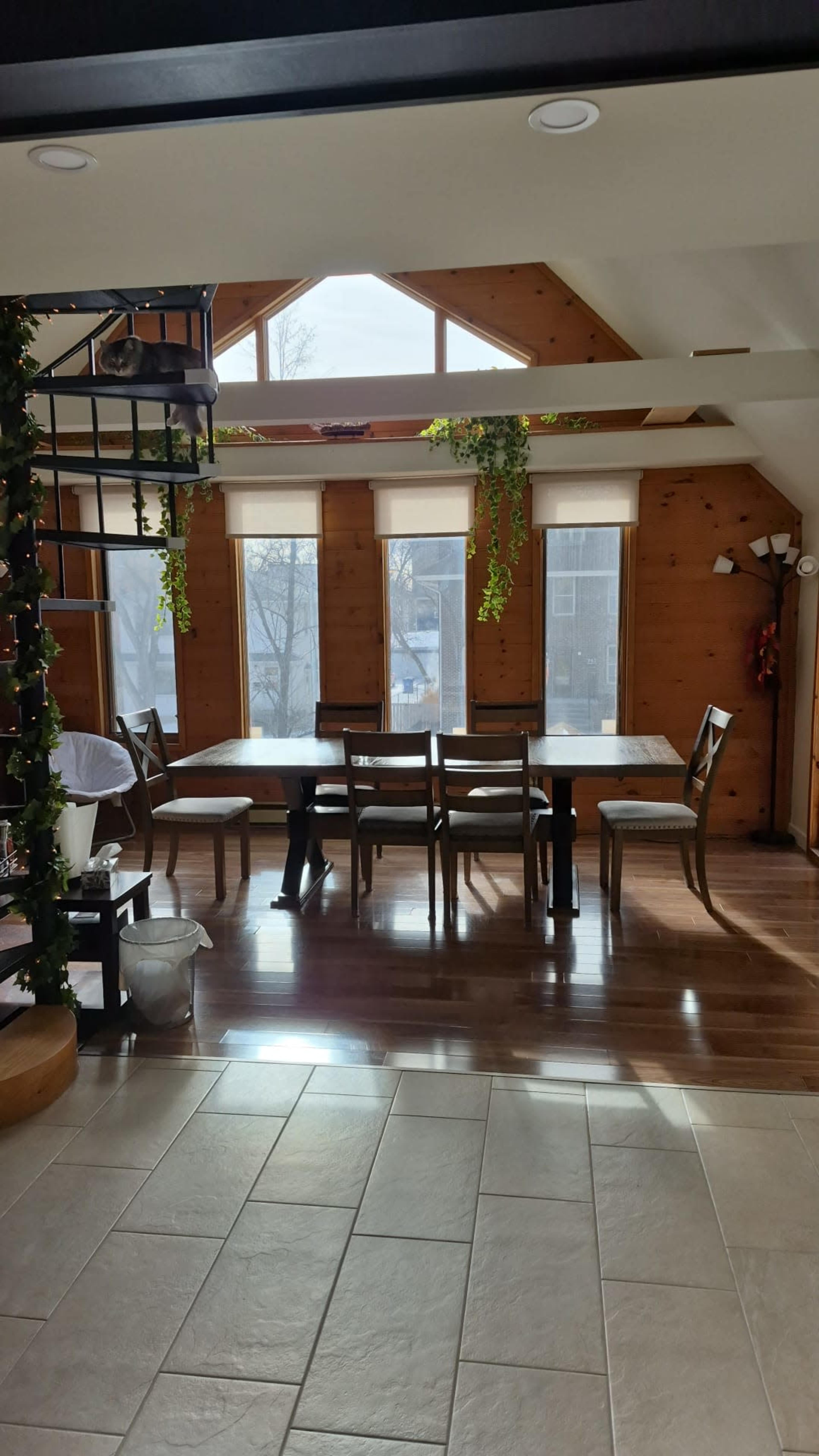 A sunlit dining area features a wooden table surrounded by chairs, with large windows and plants decorating the space.