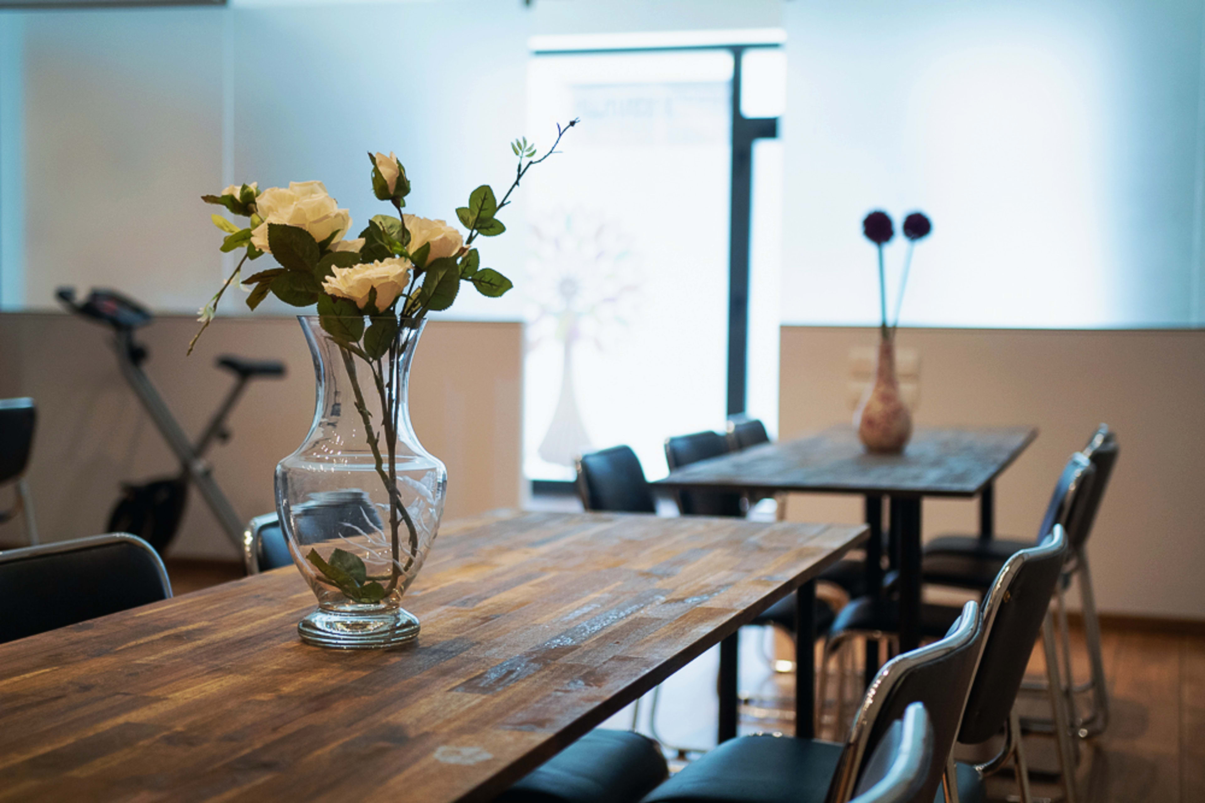 A small dining area features wooden tables with black and silver chairs, a vase of white roses, and another vase with decorative flowers, alongside a treadmill near a window.