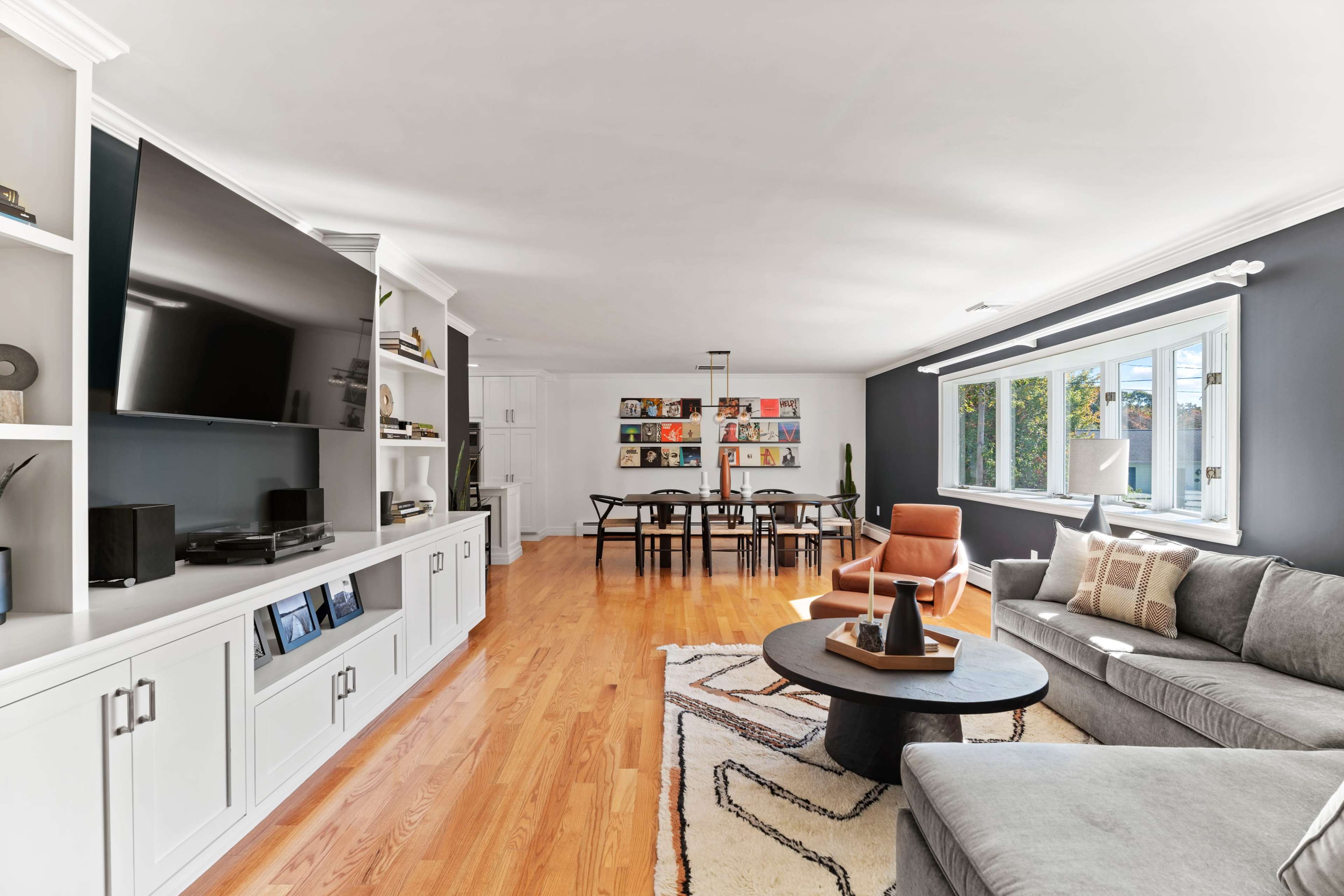 A modern living room features a large television mounted on a white cabinet, a gray sofa, an orange armchair, a round coffee table, and a dining area with a long table in the background.