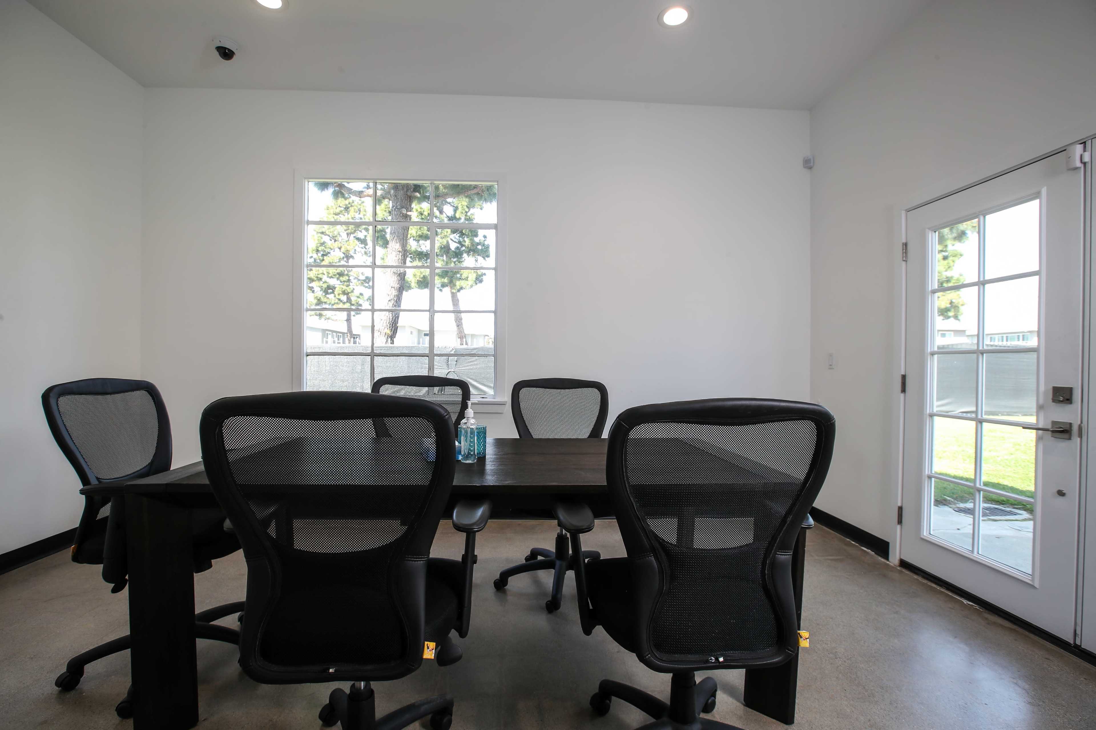 A modern conference room features a large table surrounded by five office chairs, with a window and a glass door leading to an outdoor area.