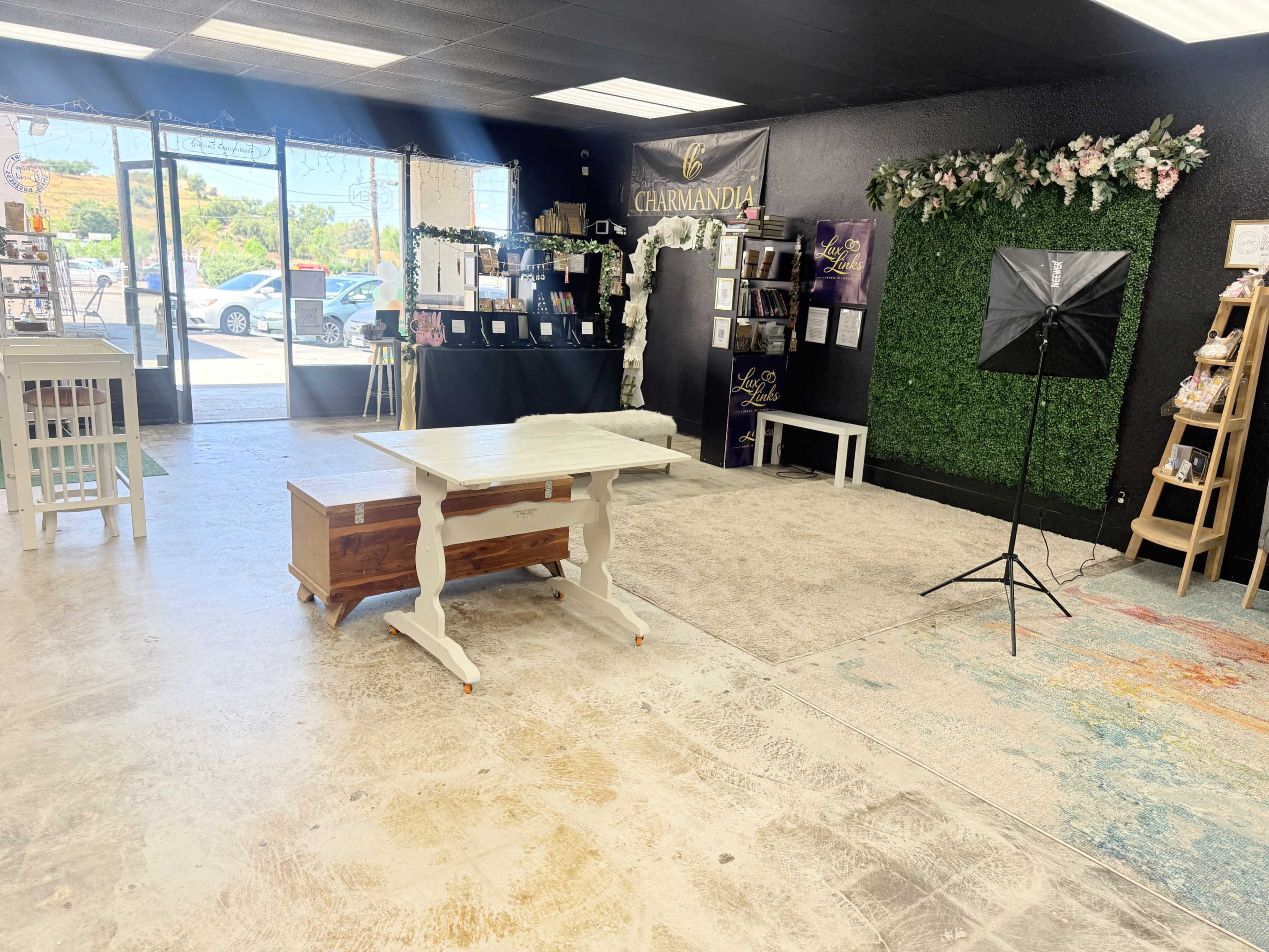 The interior of a retail space features a white wooden table, a wooden storage chest, a backdrop of green foliage, and various display shelves, with bright natural light coming through large windows.