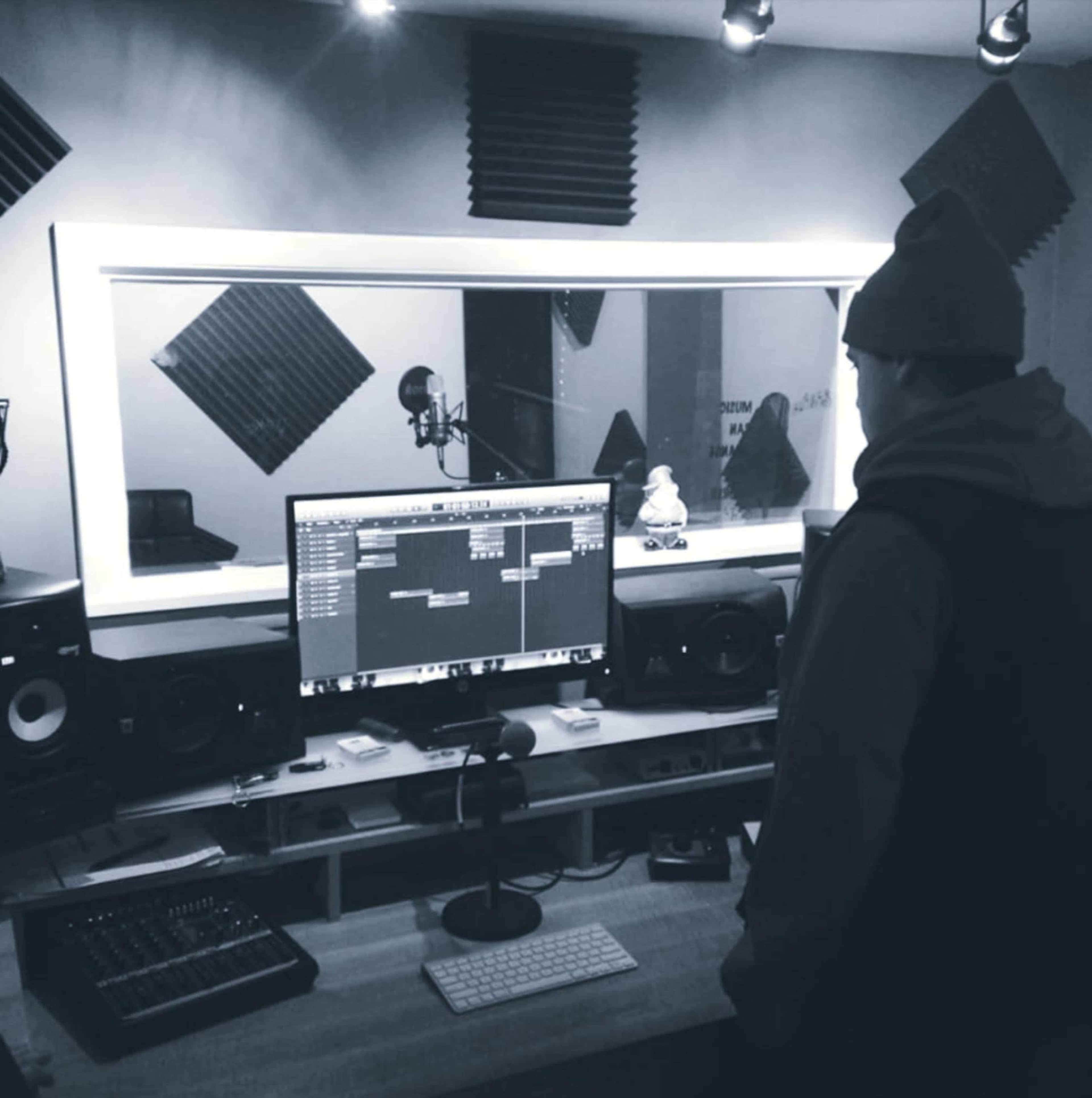 A person stands at a desk in a music studio, facing a computer displaying audio editing software, with soundproofing panels on the walls.