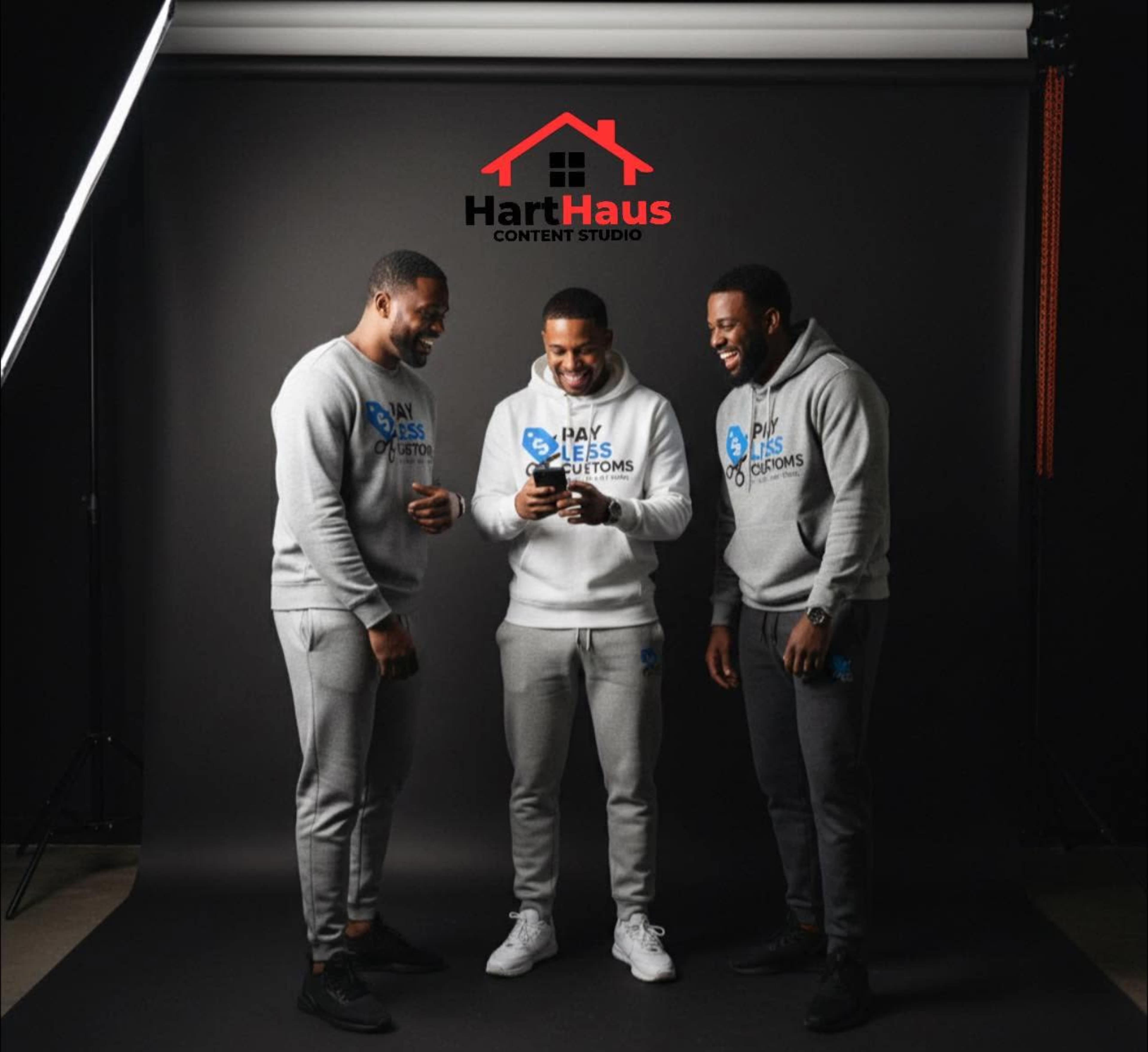 Three men in gray hoodies and sweatpants are standing in front of a black backdrop, looking at a phone together.