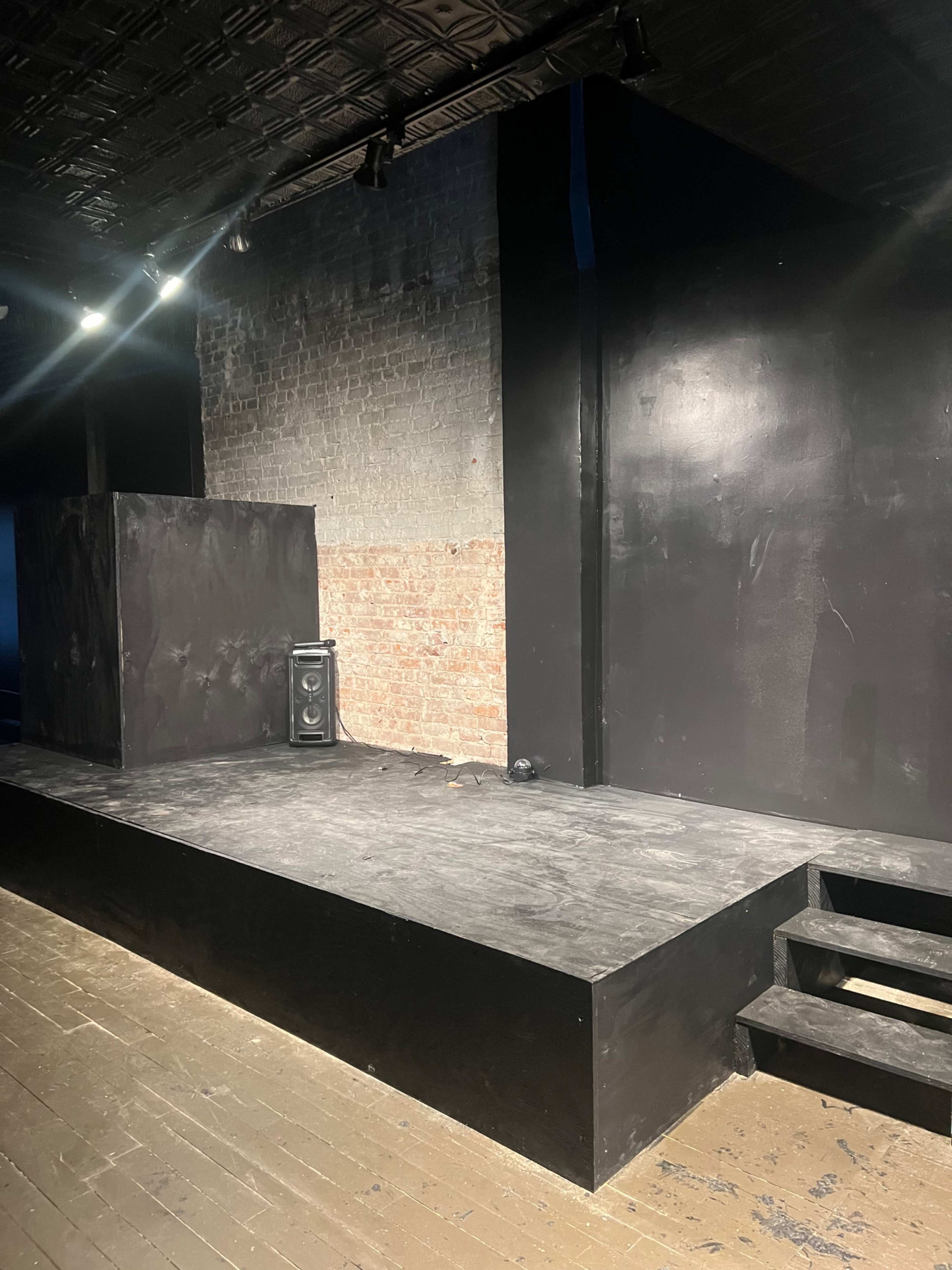 A dimly lit stage area with a black wooden platform, a brick wall in the background, and steps leading down from the stage.