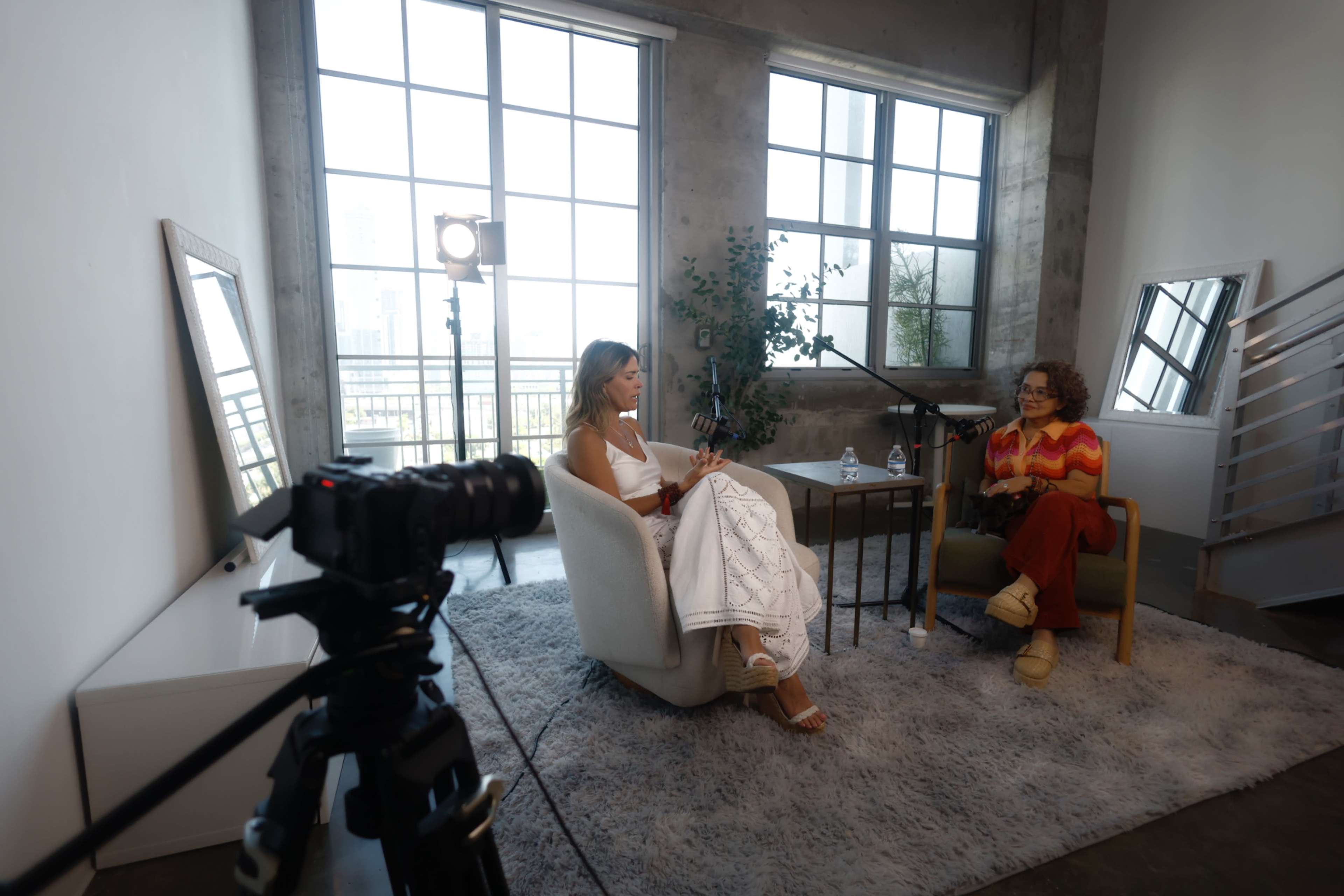 Two women are seated in an indoor setting with large windows, engaged in conversation, while a camera is positioned in the foreground.