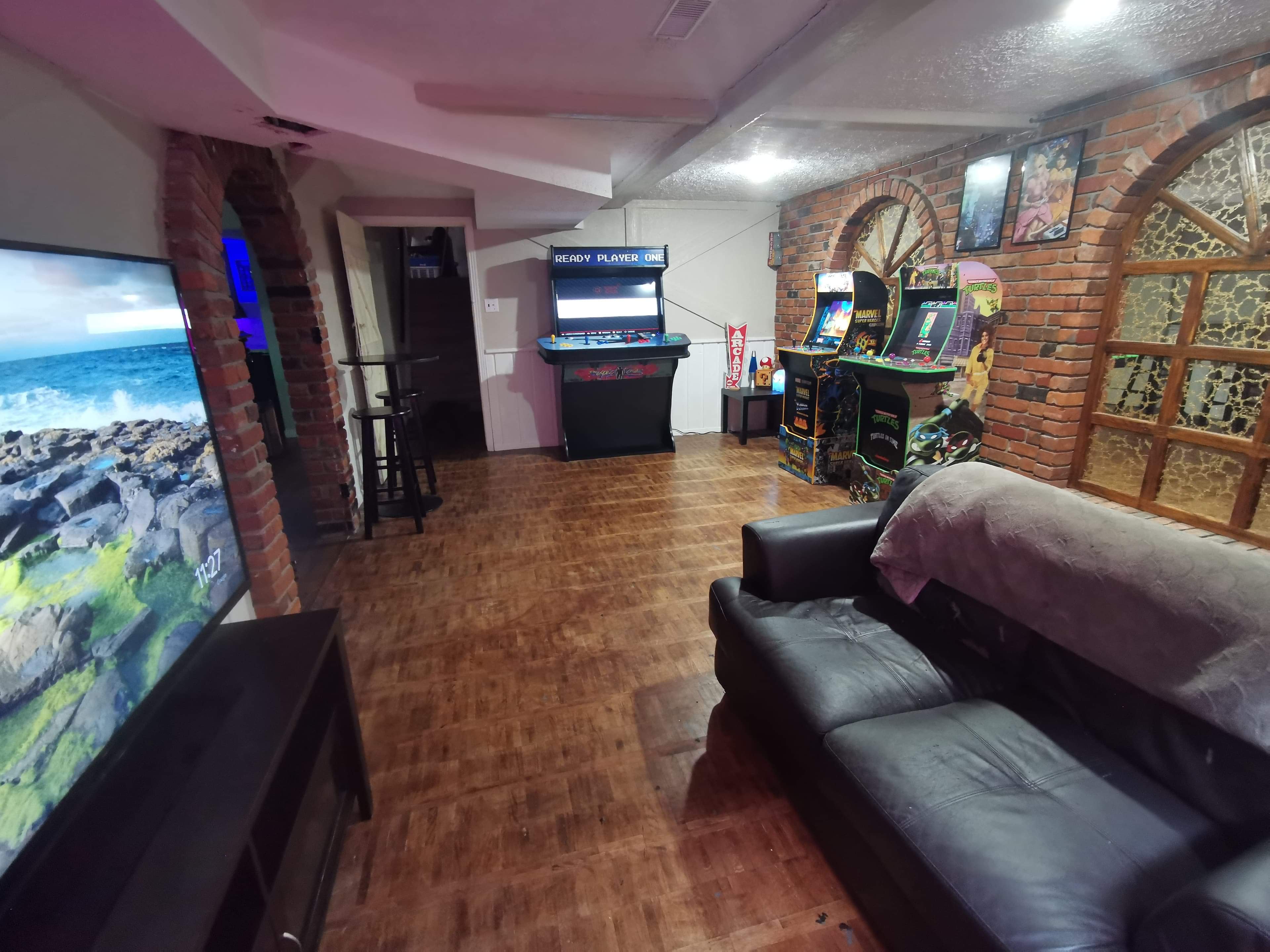 A game room with a dark leather couch, arcade machines, and a large TV set against a brick wall.