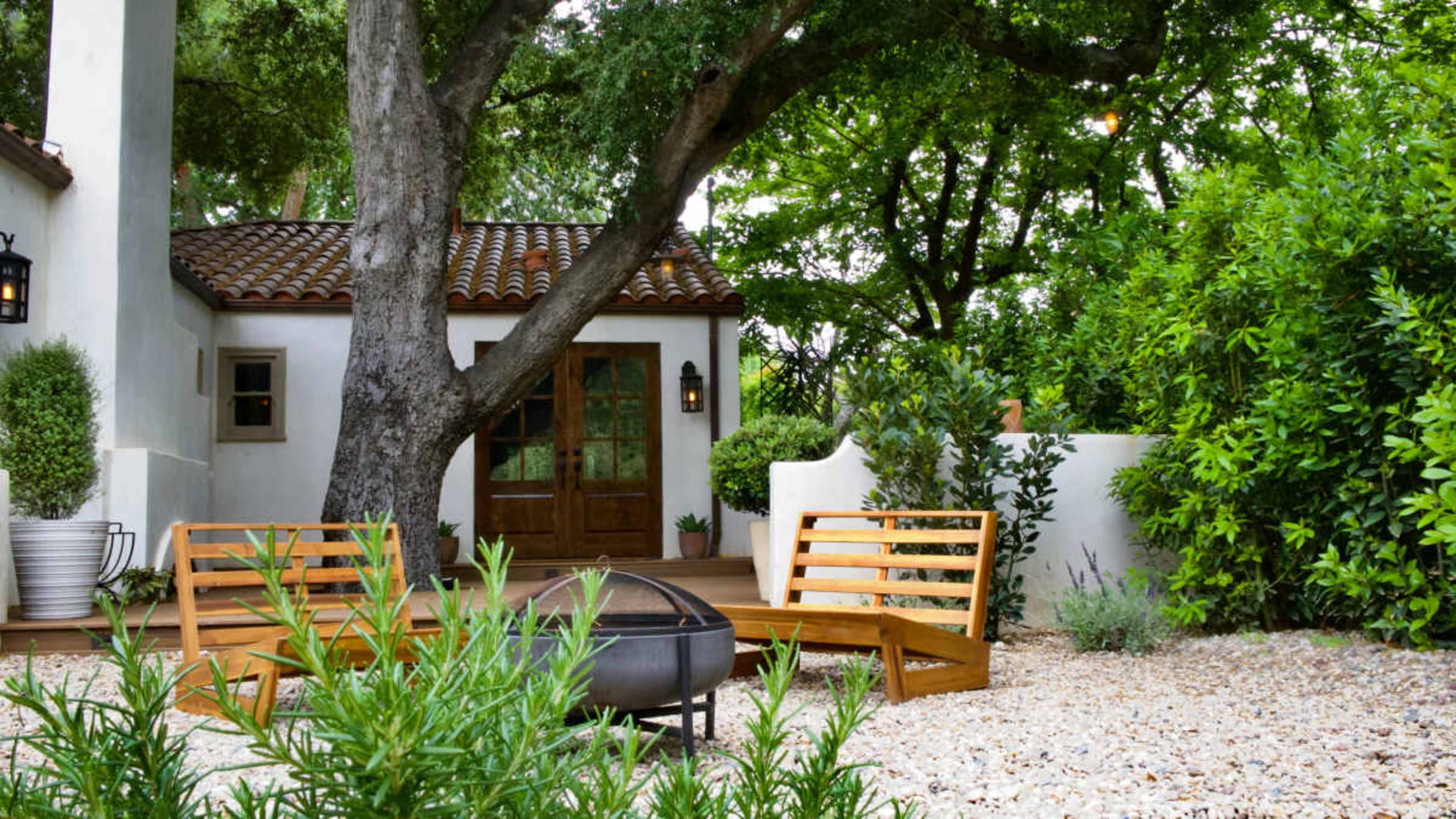 A cozy outdoor seating area with two wooden benches and a fire pit is surrounded by lush greenery and a small white building.