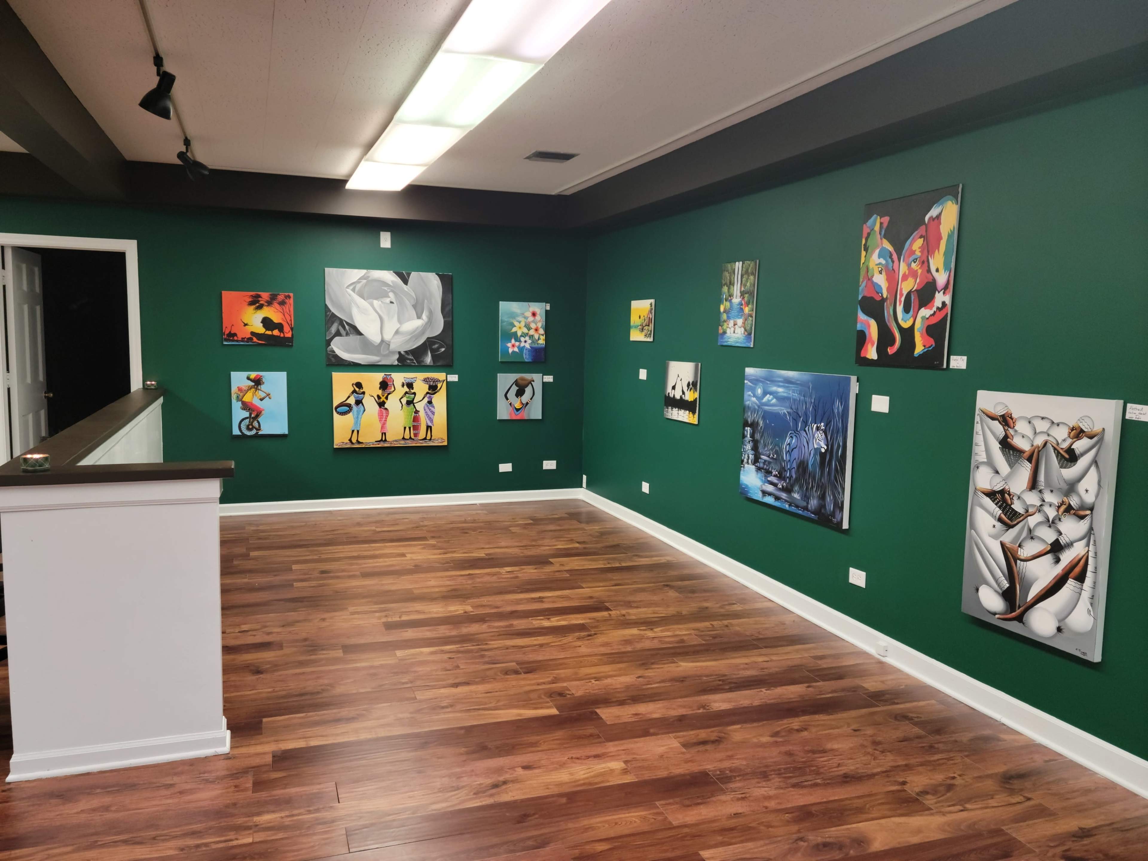 A gallery interior with green walls displaying various colorful paintings and a wooden floor.