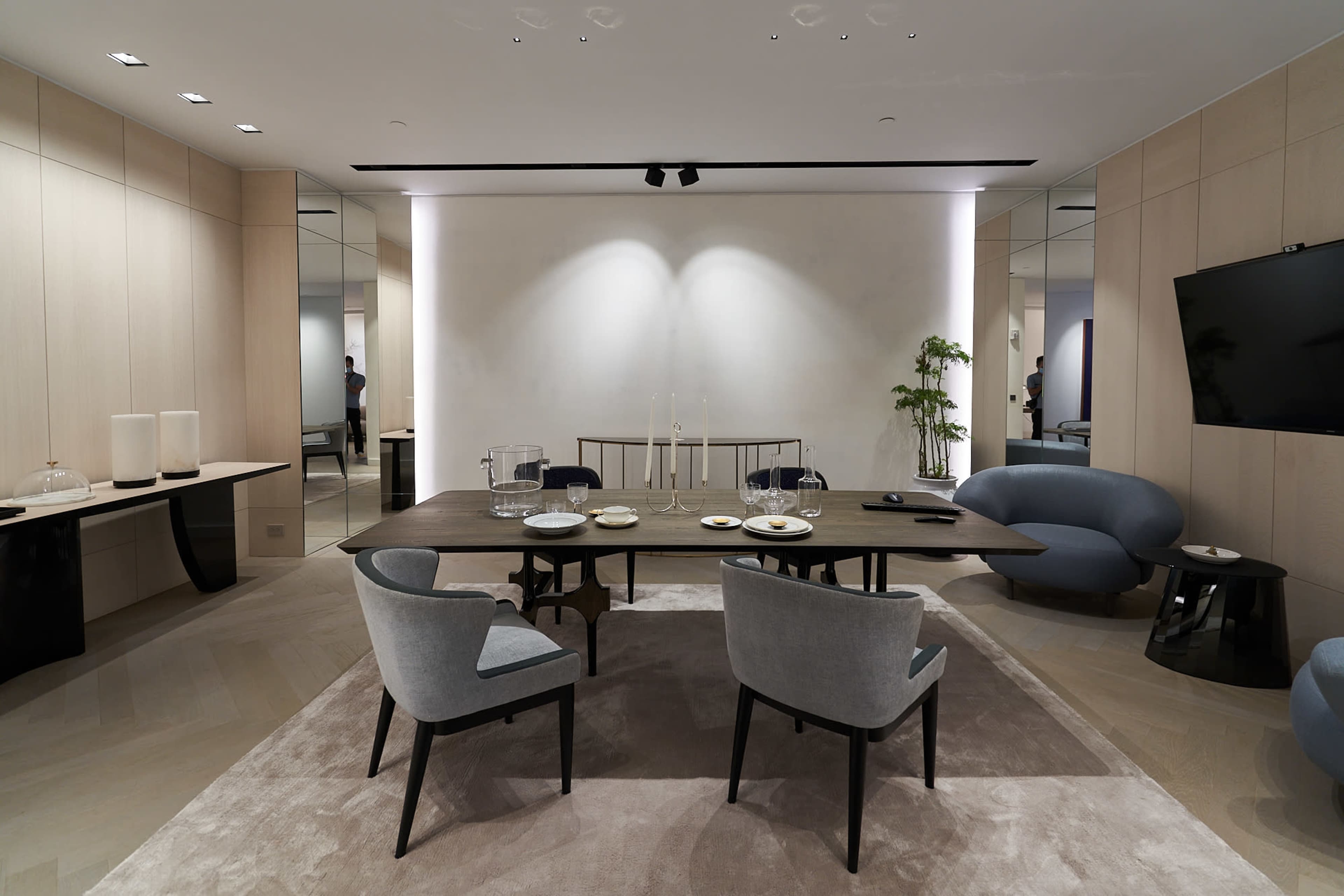 A modern dining area features a rectangular table surrounded by four chairs on a light rug, with minimalist decor and wall-mounted TV in the background.
