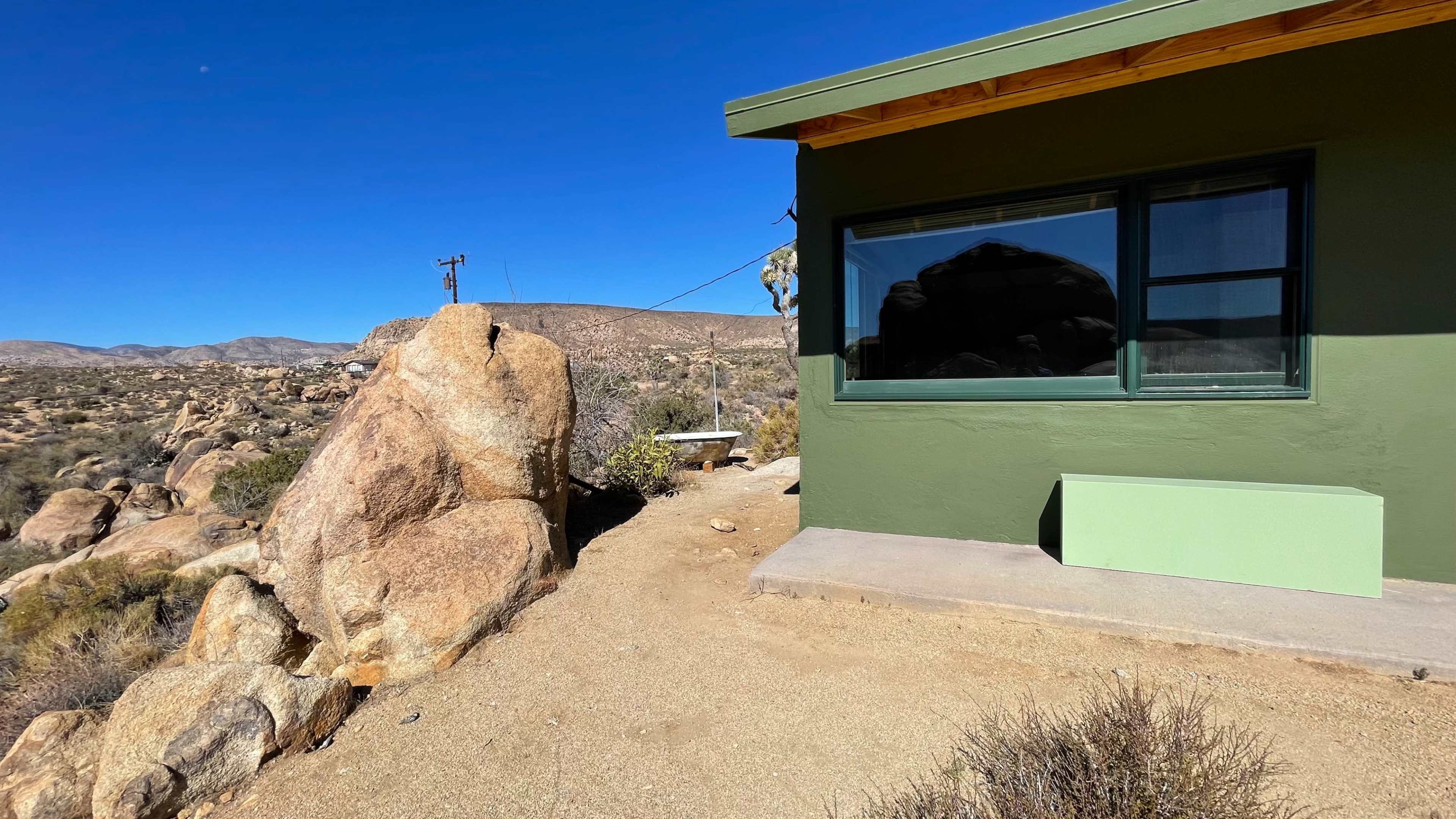 A large boulder stands near a green building in a desert landscape.