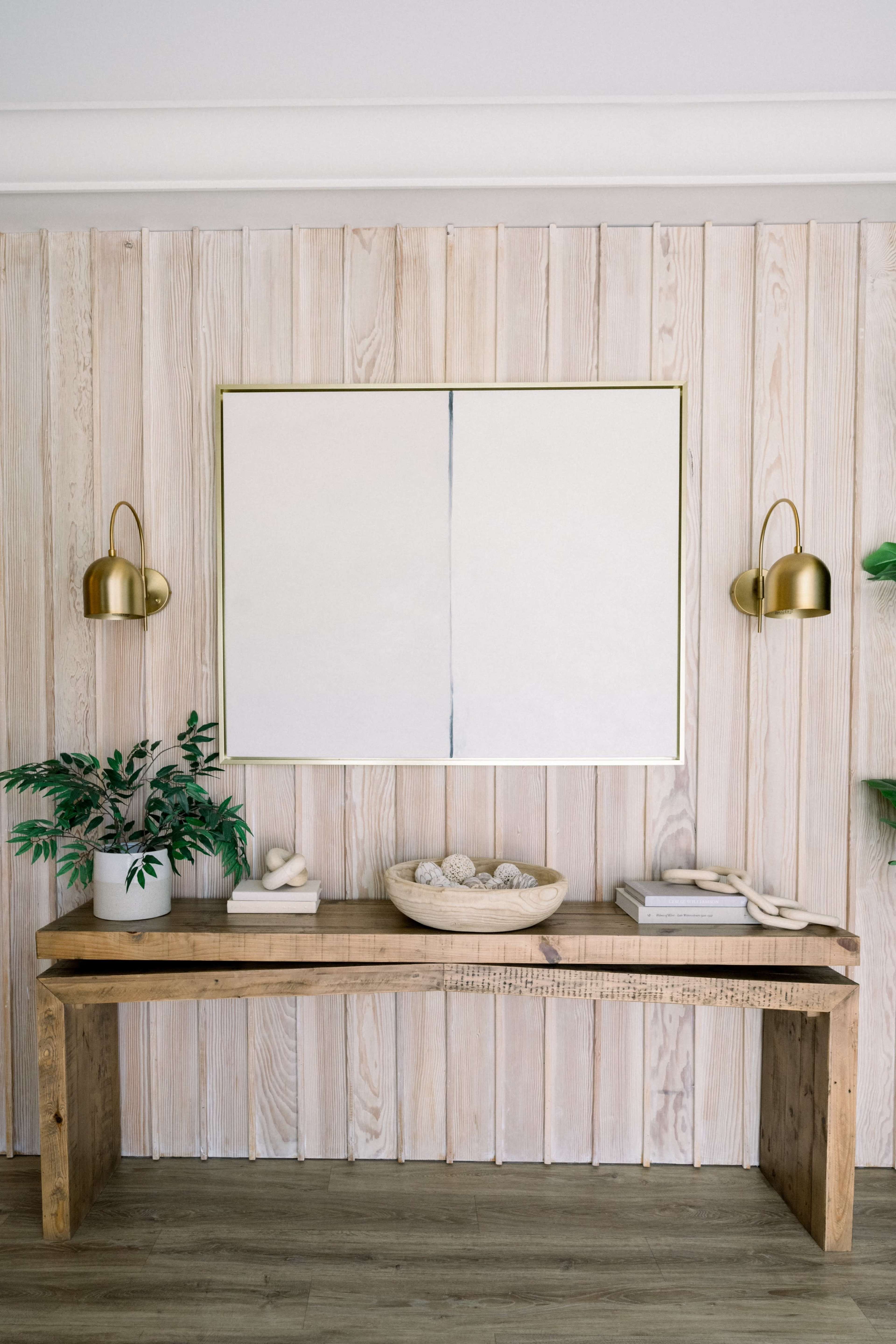 A wooden console table is positioned against a light wooden panel wall, featuring a large blank framed artwork and two brass wall sconces, accompanied by a plant and decorative items on the table.