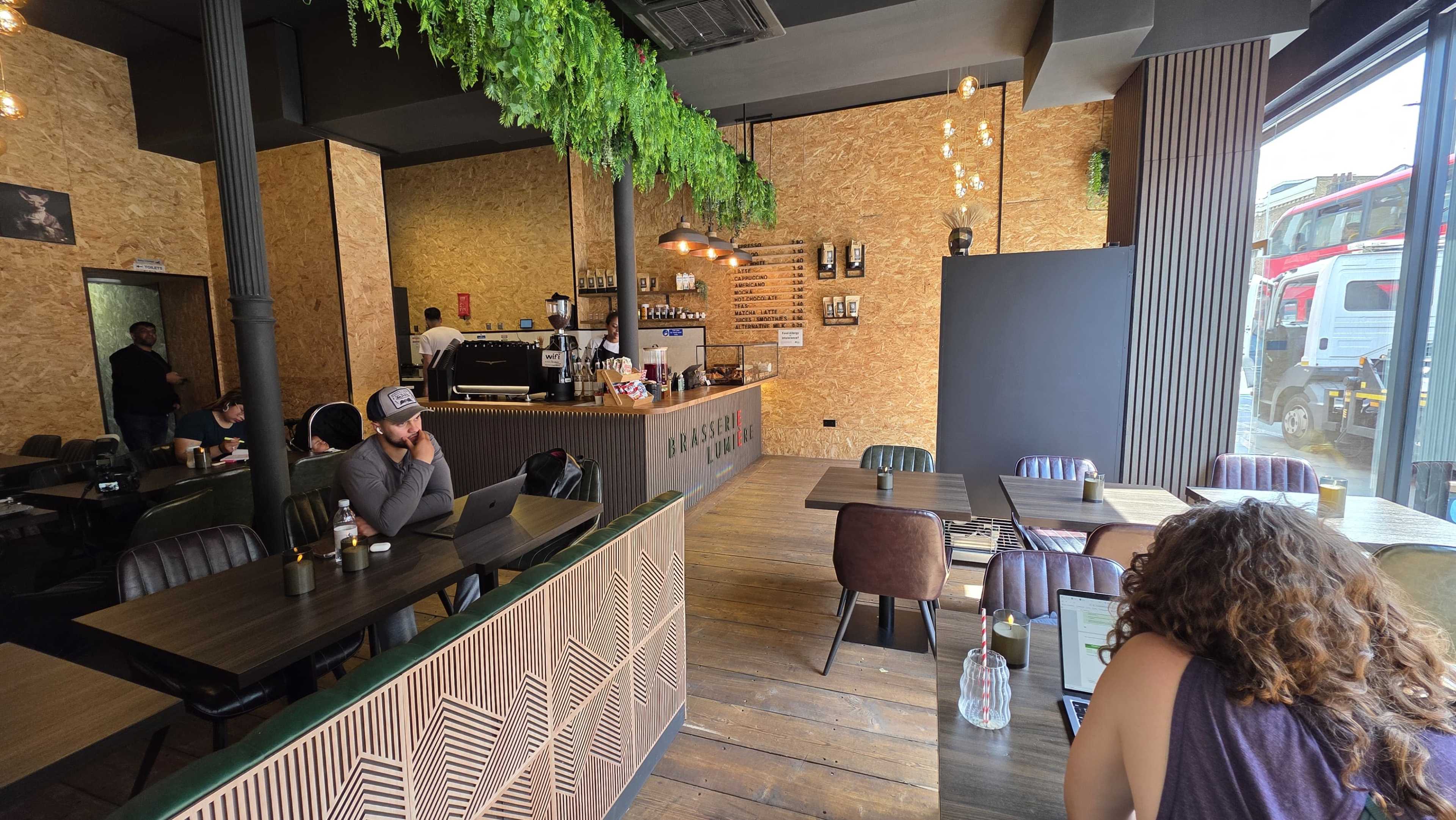 The image shows a modern café interior with wooden flooring, green plants hanging from the ceiling, and patrons seated at tables, while a coffee counter is visible in the background.