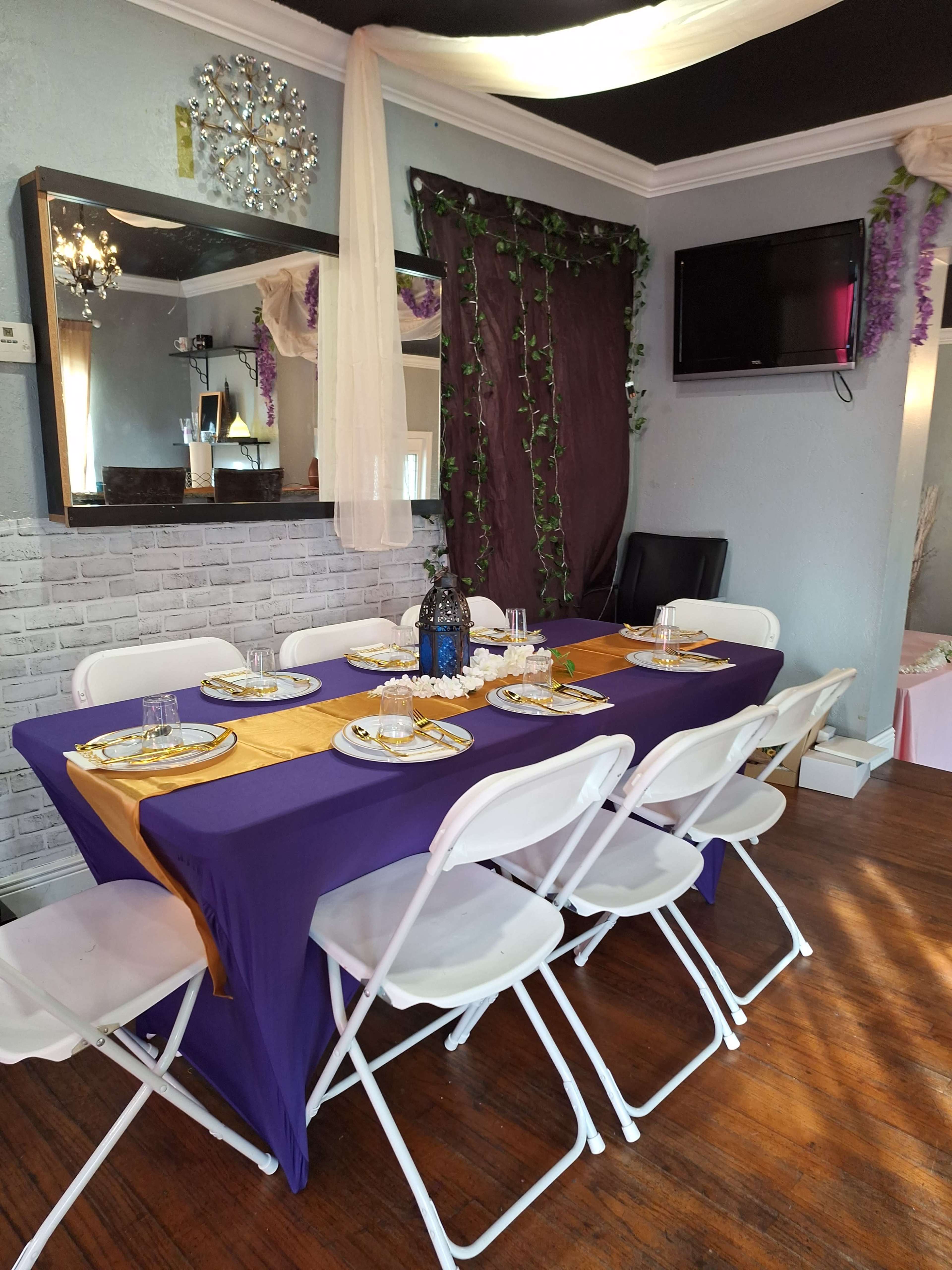 A dining table set with a purple tablecloth and gold accents is arranged with white chairs in a decorated indoor space.