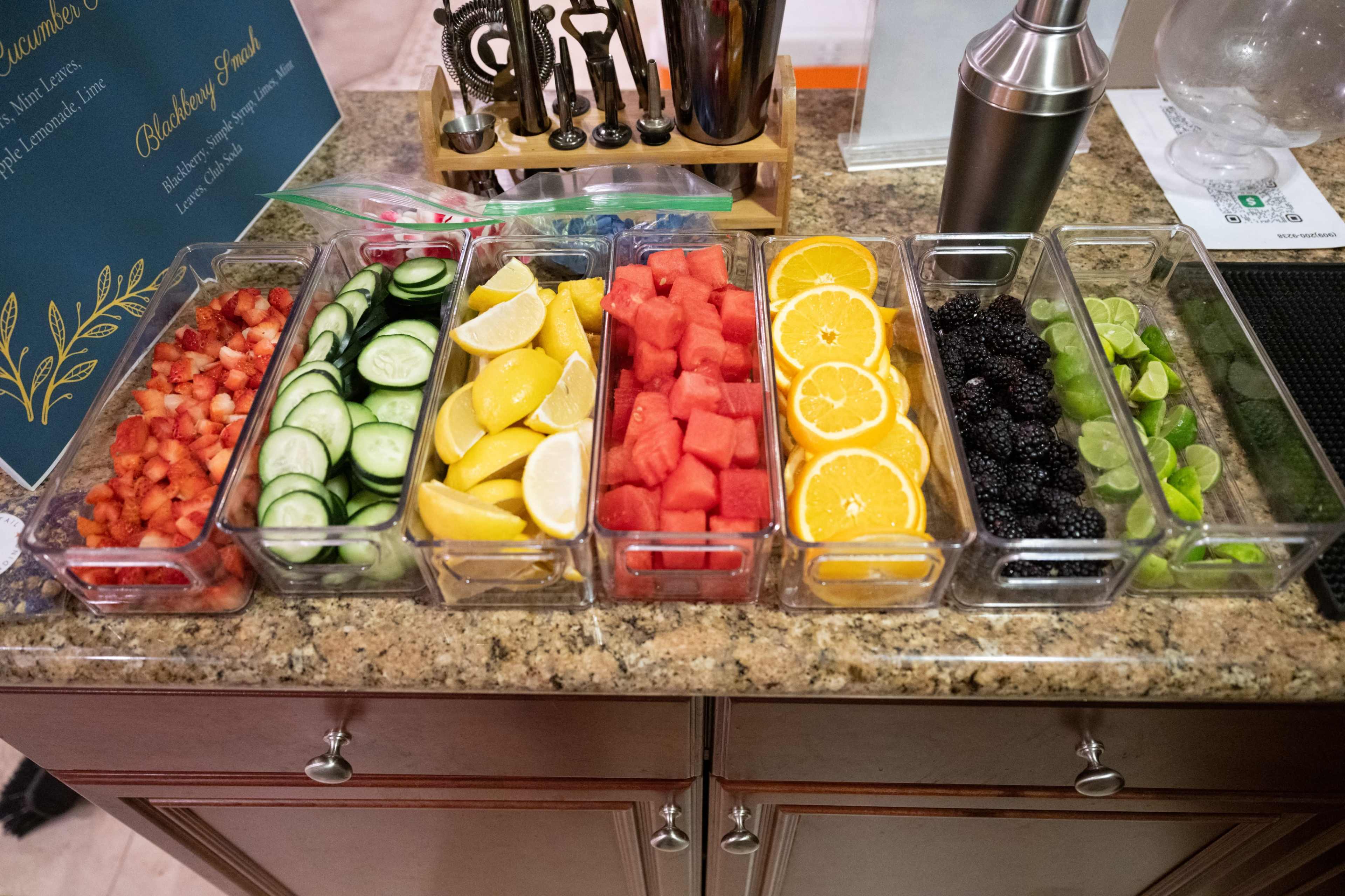 A countertop with six clear containers filled with various sliced fruits and vegetables, including strawberries, cucumbers, lemons, watermelon, oranges, and blackberries.