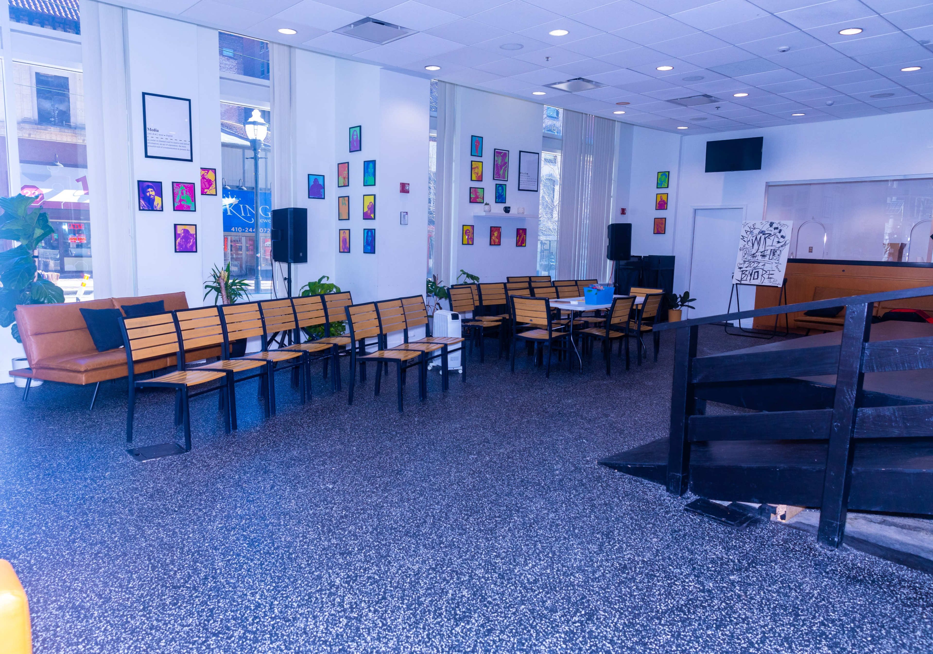The image shows a spacious, modern interior with rows of wooden benches, a few potted plants, and colorful artwork displayed on the walls.
