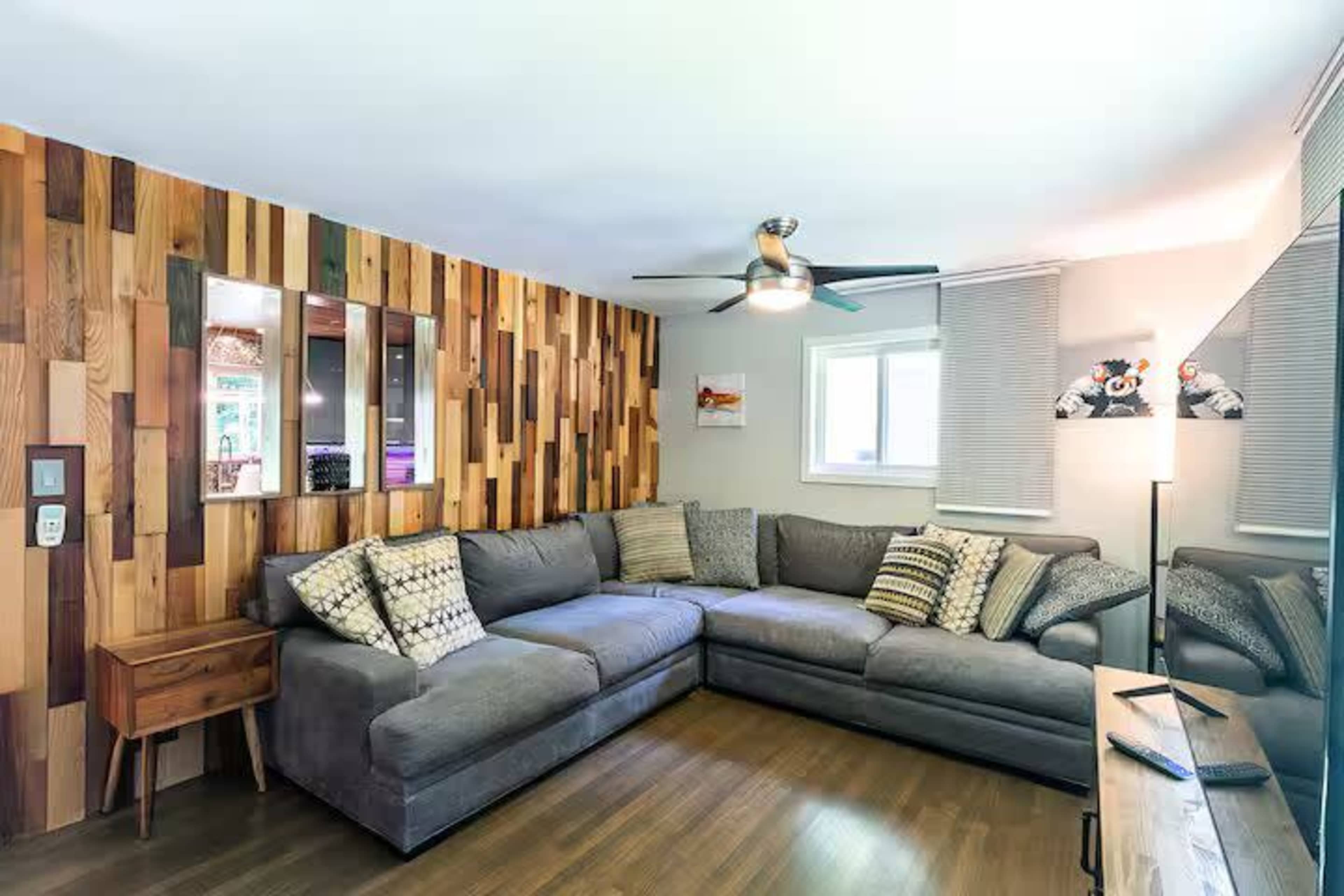 The image shows a modern living room with a sectional sofa, wooden accent wall, and a ceiling fan.