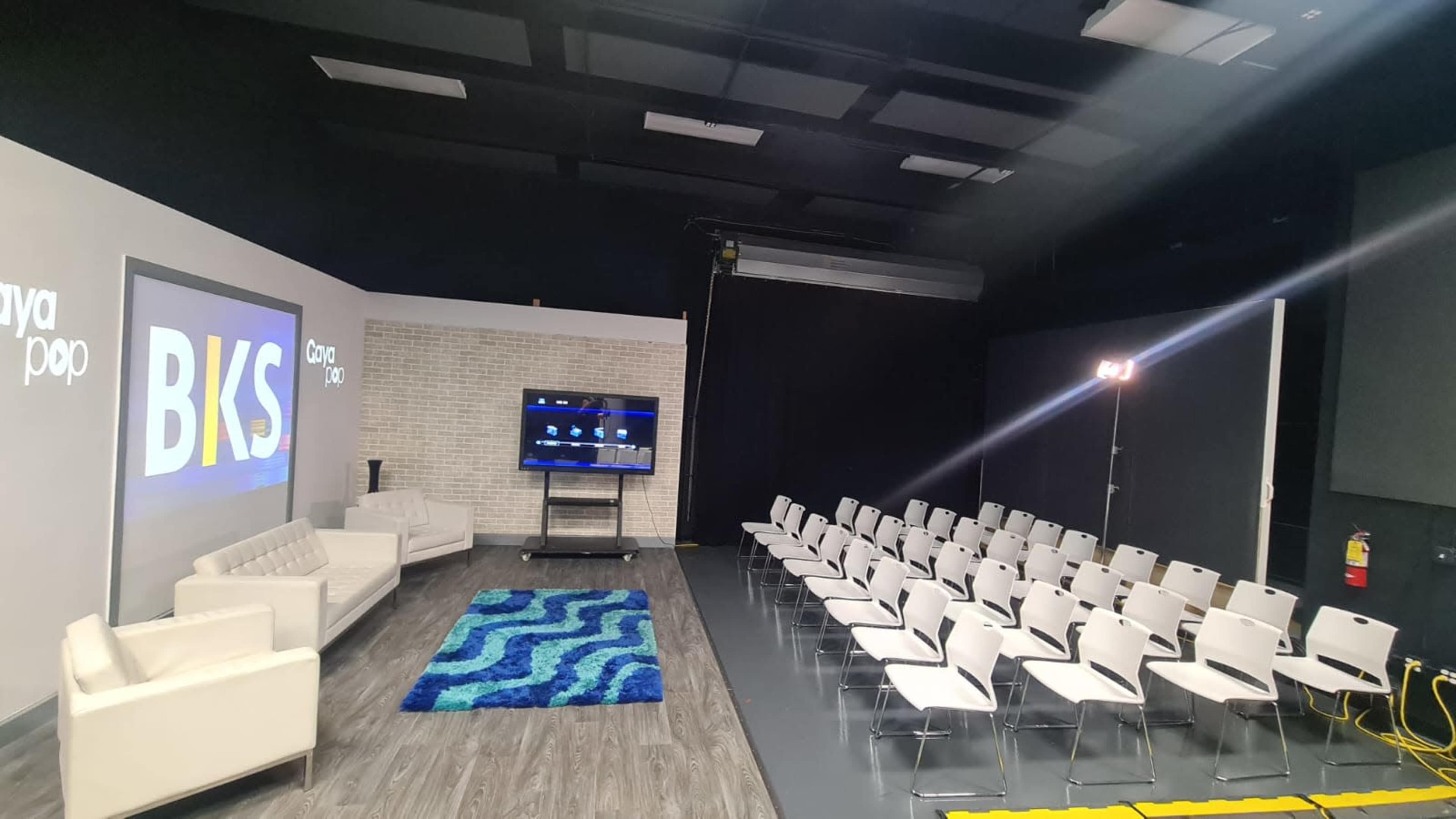 The image shows a modern studio setup with white chairs arranged in rows facing a stage, featuring a blue rug, two white sofas, and a television display.