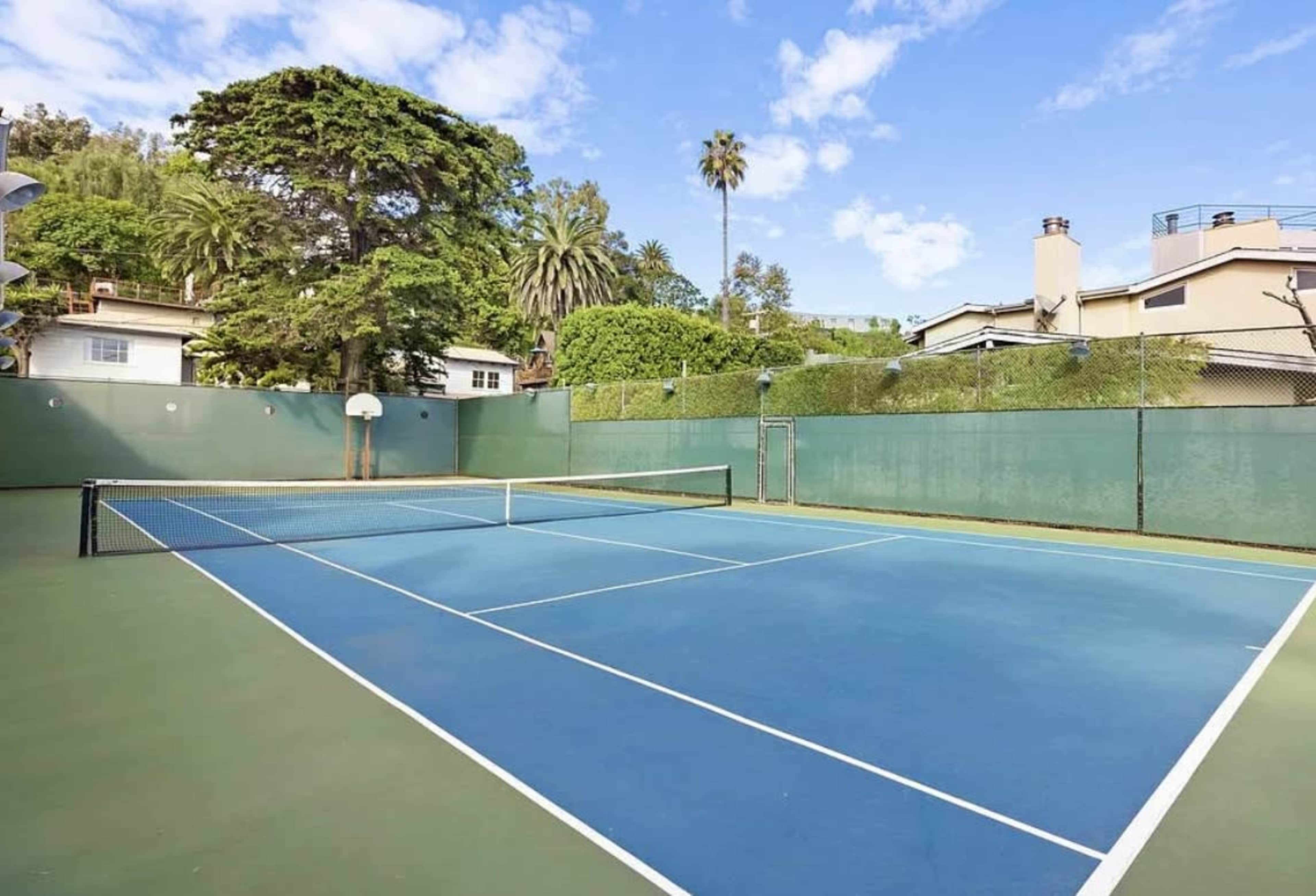 The image shows an outdoor tennis court surrounded by greenery and residential buildings under a partly cloudy sky.