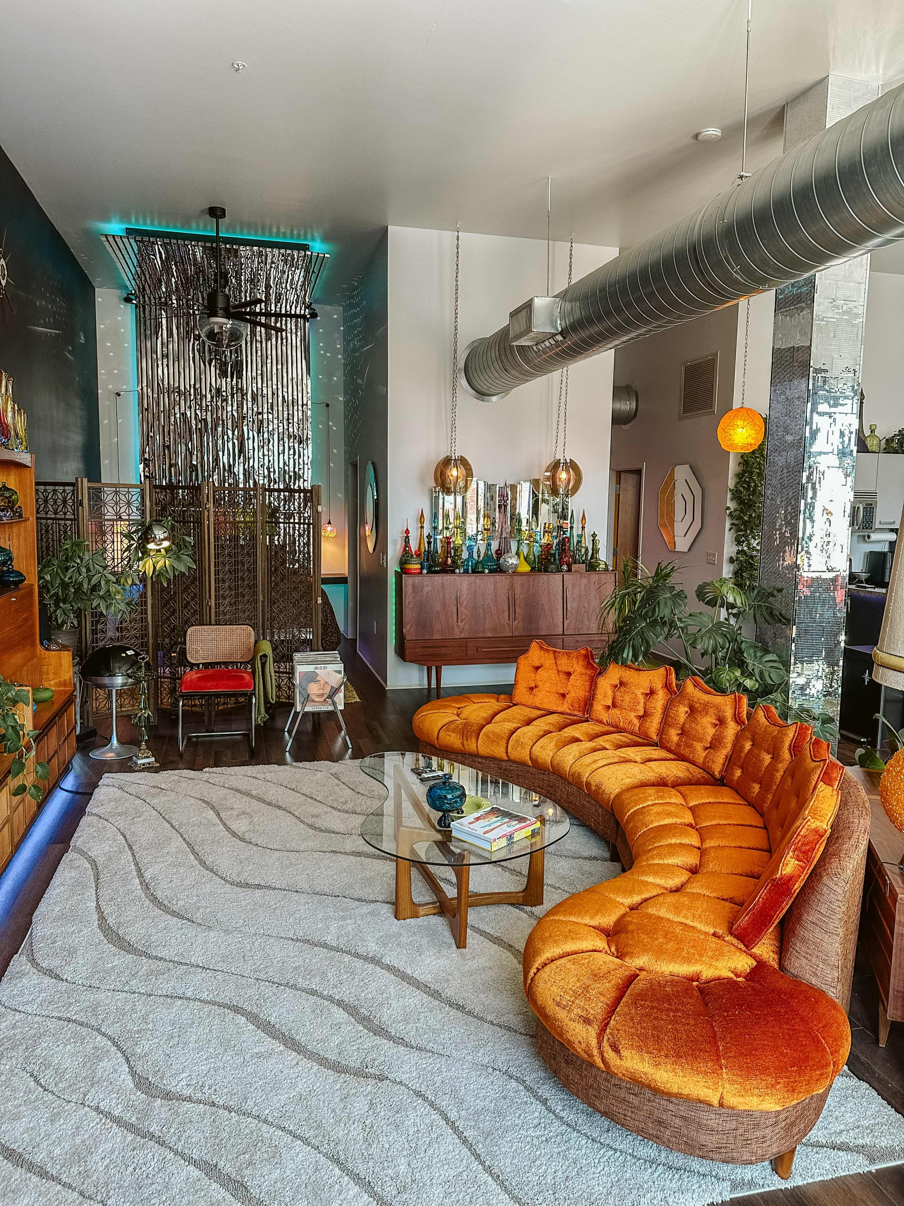The image depicts a modern living room featuring an orange curved sofa, a glass coffee table, and plants, with a backdrop of decorative shelves filled with colorful bottles.