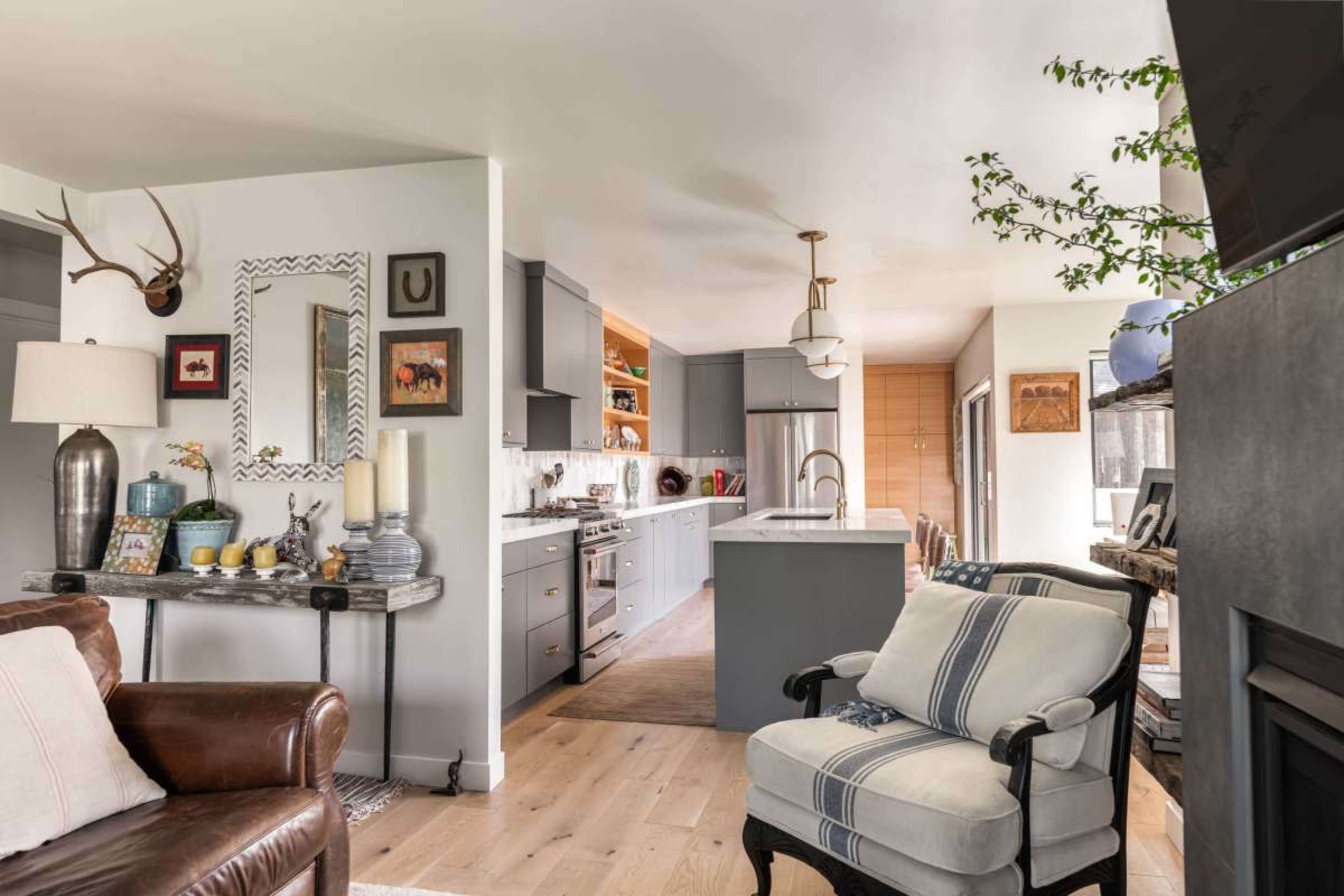 The image shows a modern kitchen with gray cabinetry, a large island, and an open layout that connects to a cozy living area featuring a leather chair and decorative accents.