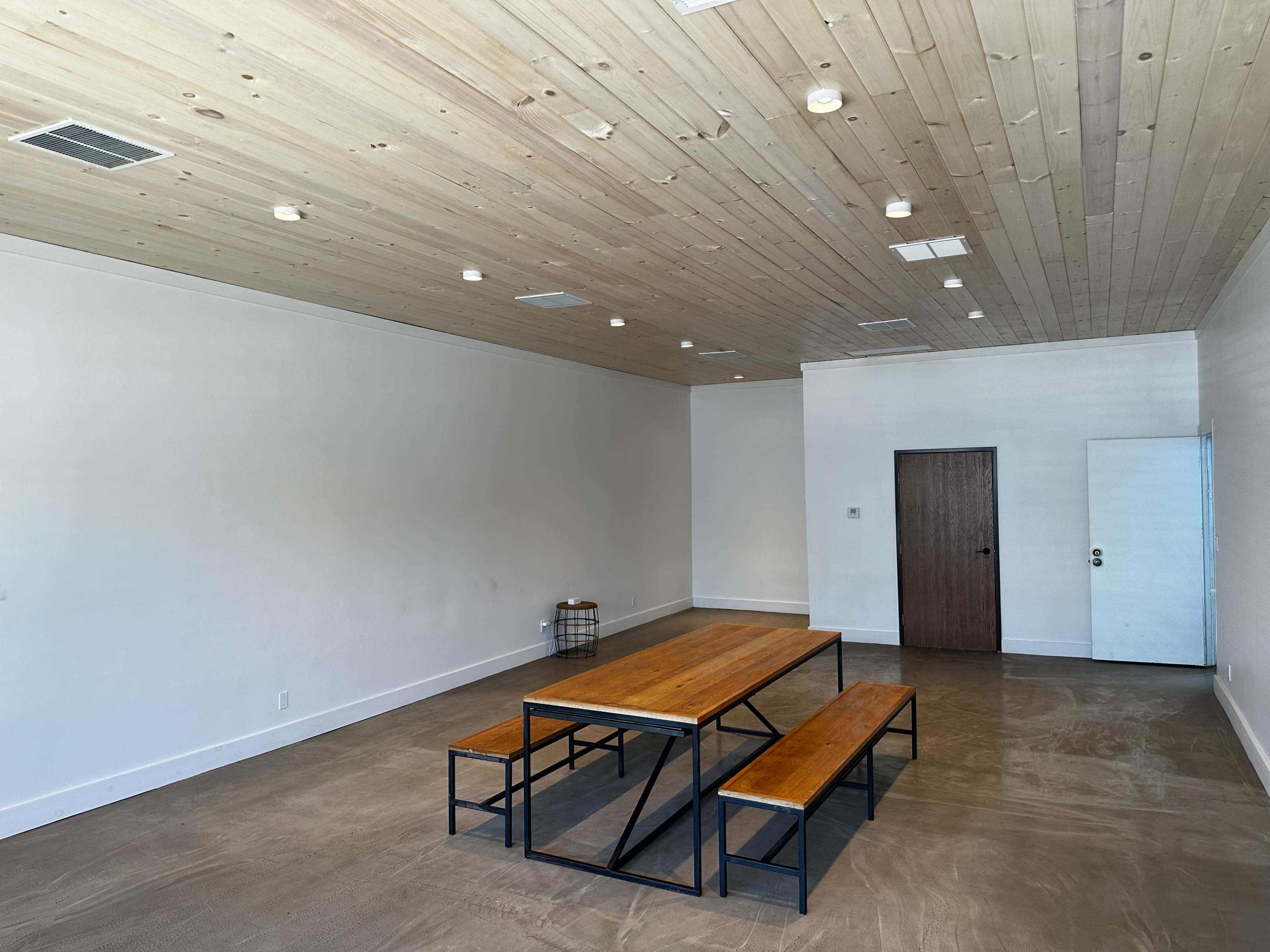A spacious room with a wooden ceiling, a metal-framed table, and benches made of wood, set on a concrete floor.