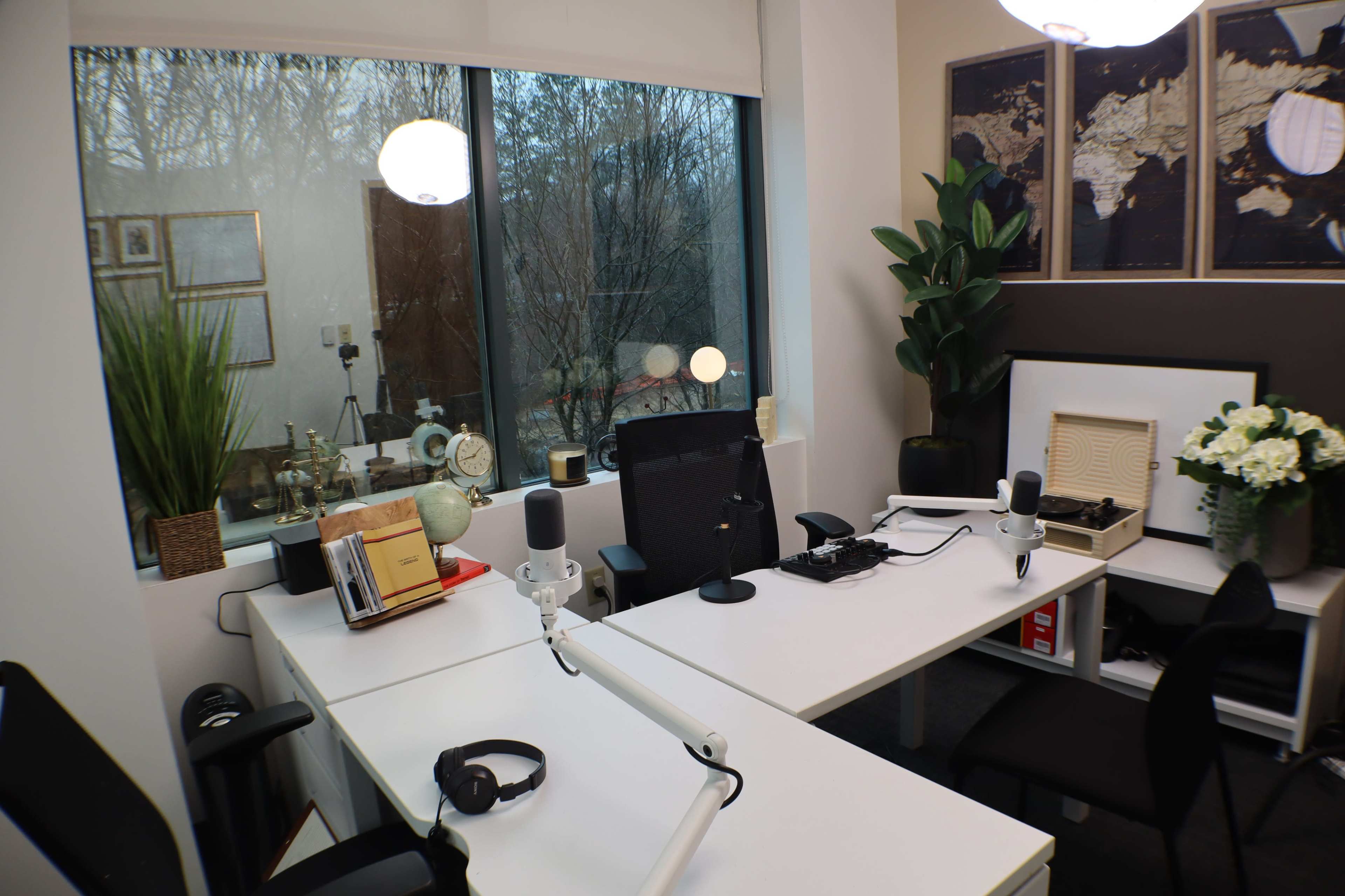 The image shows a modern office space designed for a podcast, featuring two desks with microphones, headphones, and decorative plants, located near large windows.