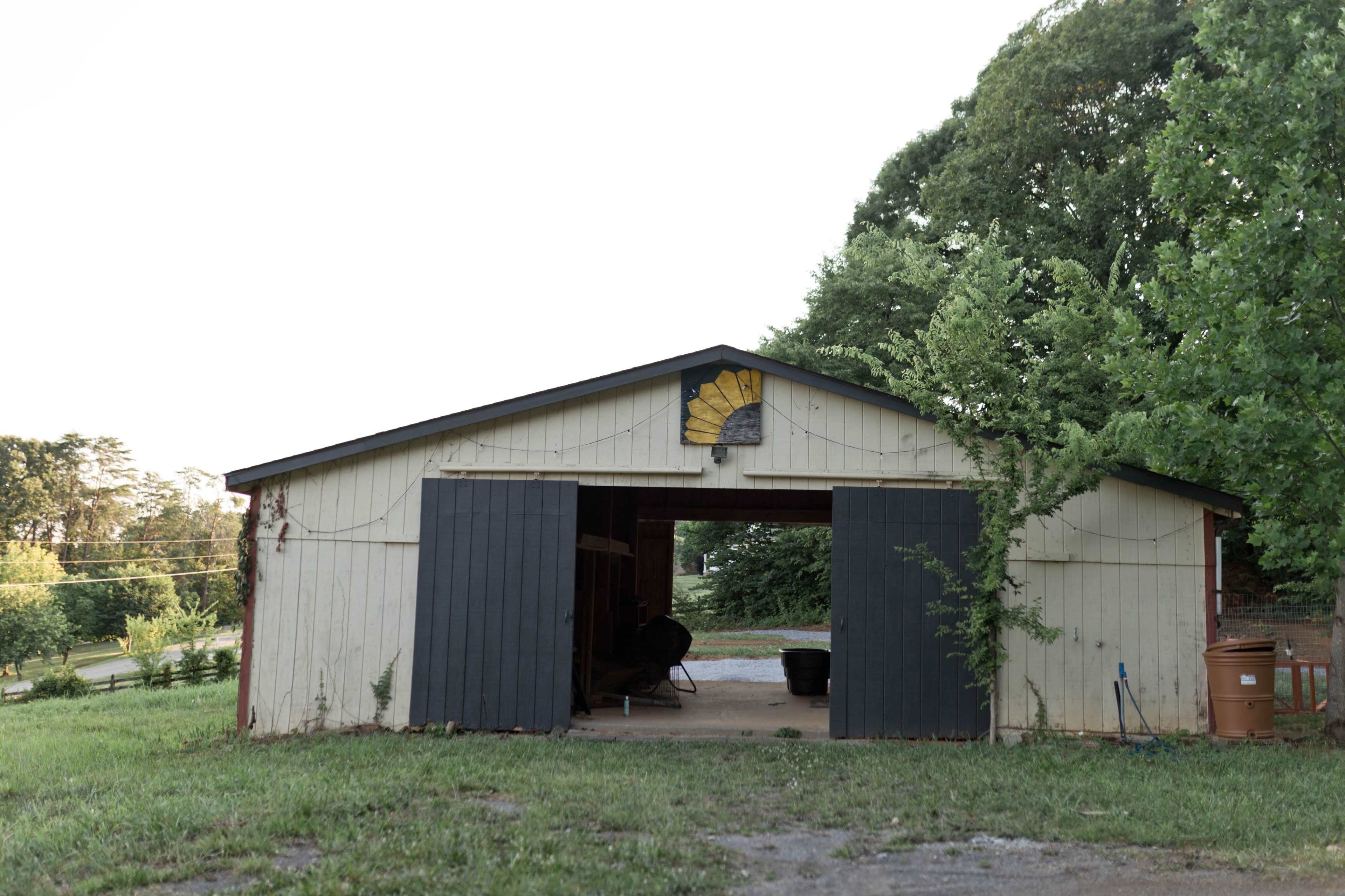 The image shows a weathered barn with dark blue doors, situated on a grassy area surrounded by trees.