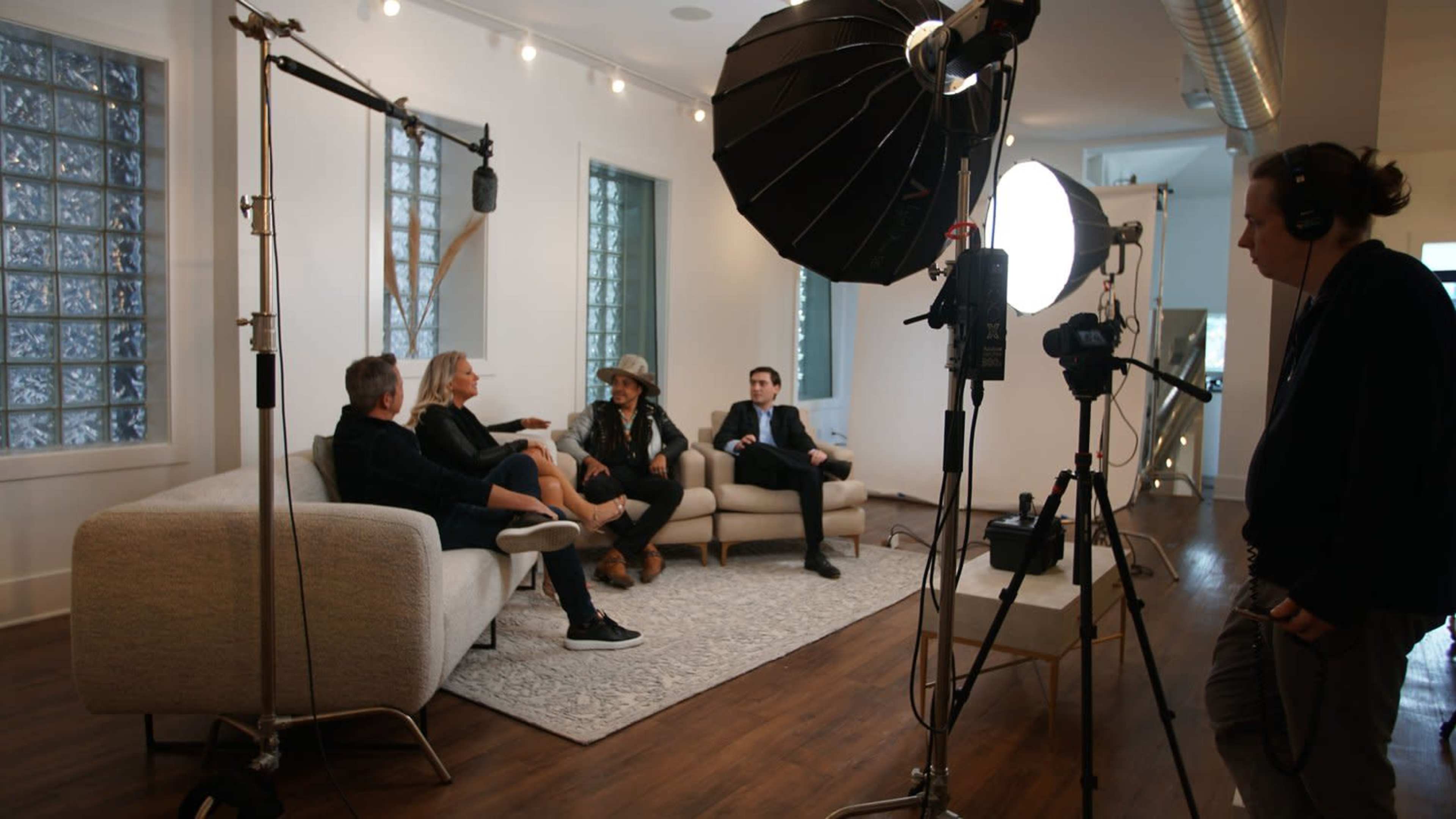 A film crew is recording an interview with four people seated on a couch in a well-lit room with modern decor.