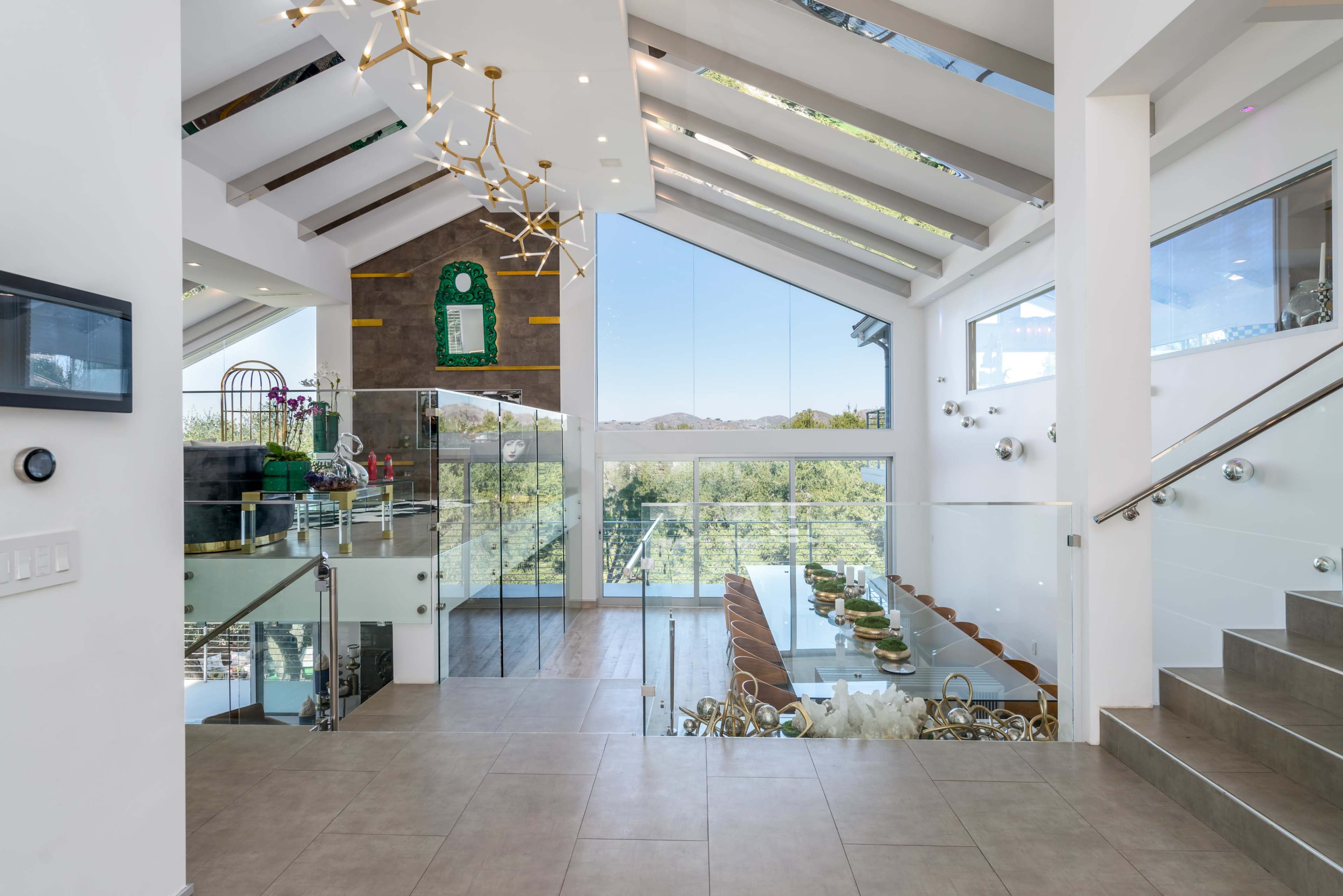 A modern open-concept living space featuring a glass railing staircase, a long dining table, and large windows that provide views of the outdoors.