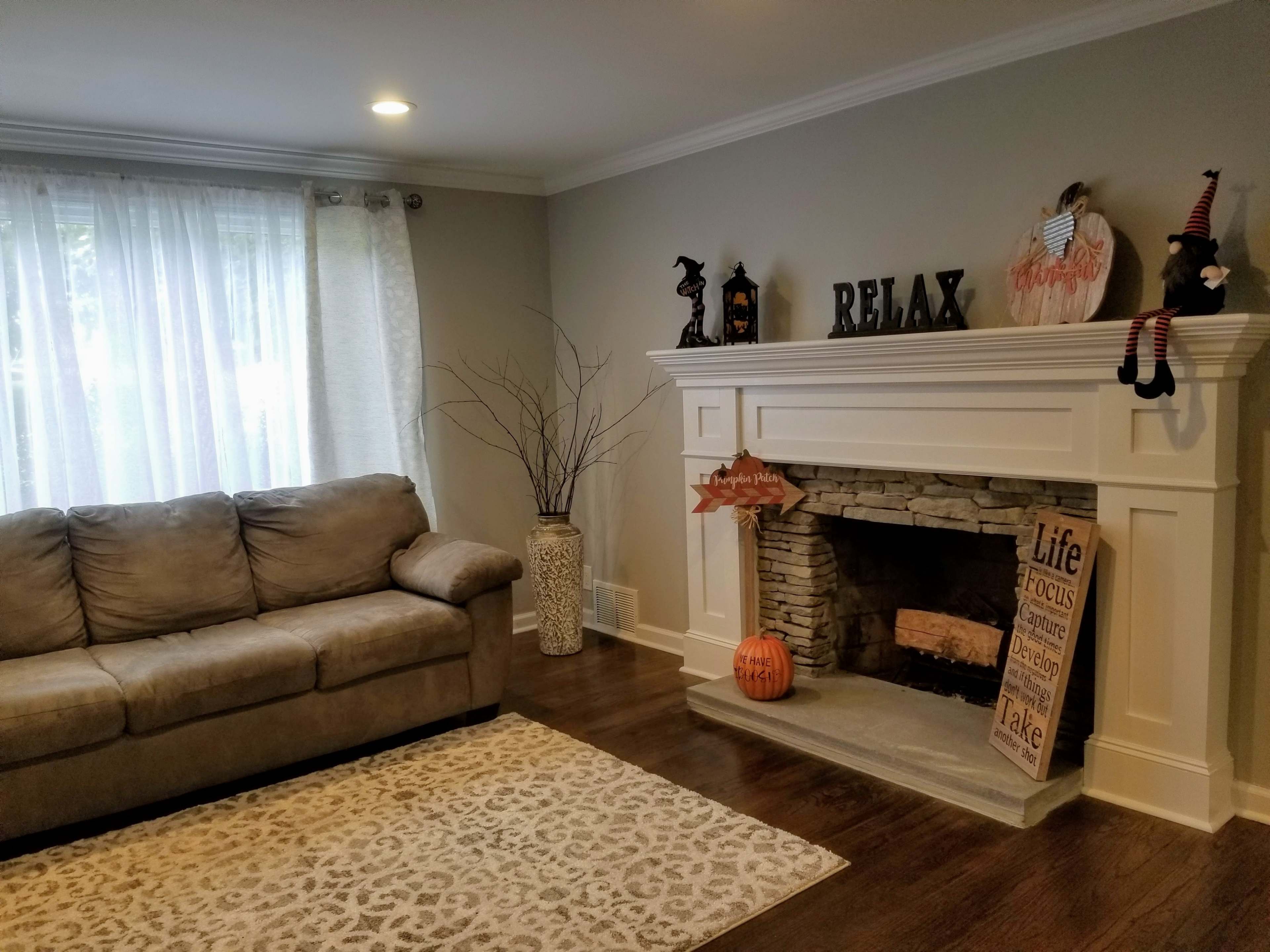 A living room features a gray sofa, a stone fireplace with seasonal decorations, and a large window with sheer curtains.