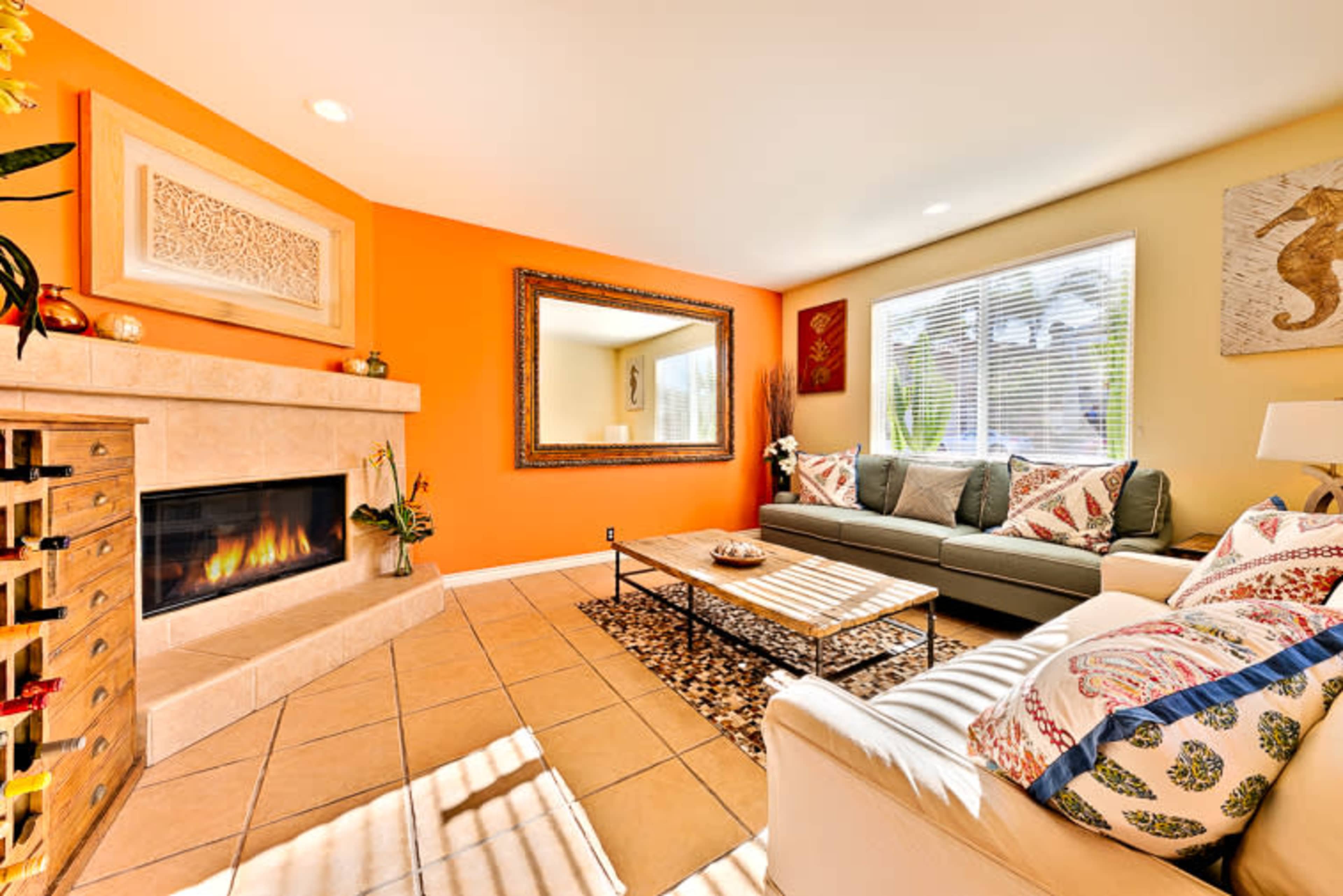 A living room features a beige sofa, a grey couch, a coffee table, and a fireplace, with an orange accent wall and ample natural light from the windows.