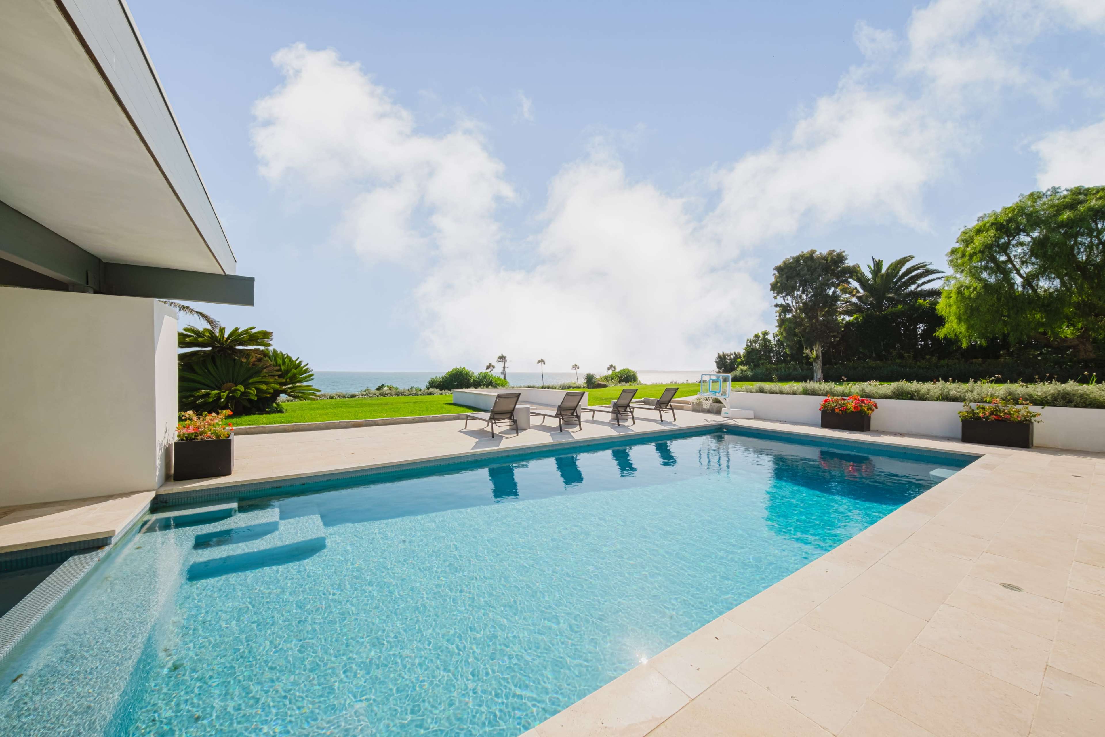 The image shows a streamlined pool area with lounge chairs overlooking a green landscape and the ocean under a bright sky.