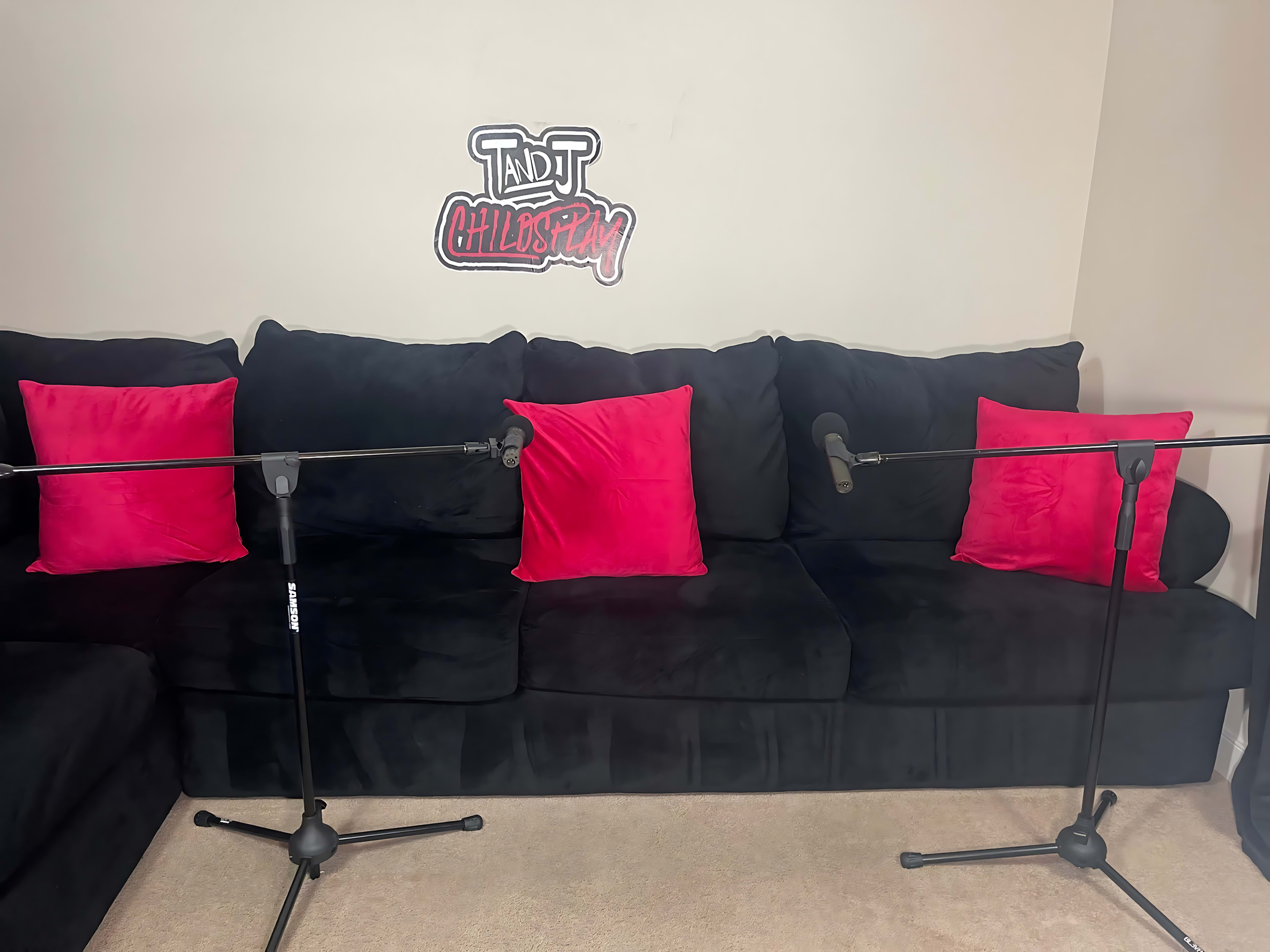A black couch with two pink pillows and two microphone stands positioned in front of it, against a wall featuring a sign that reads "T and T Chill and Play."