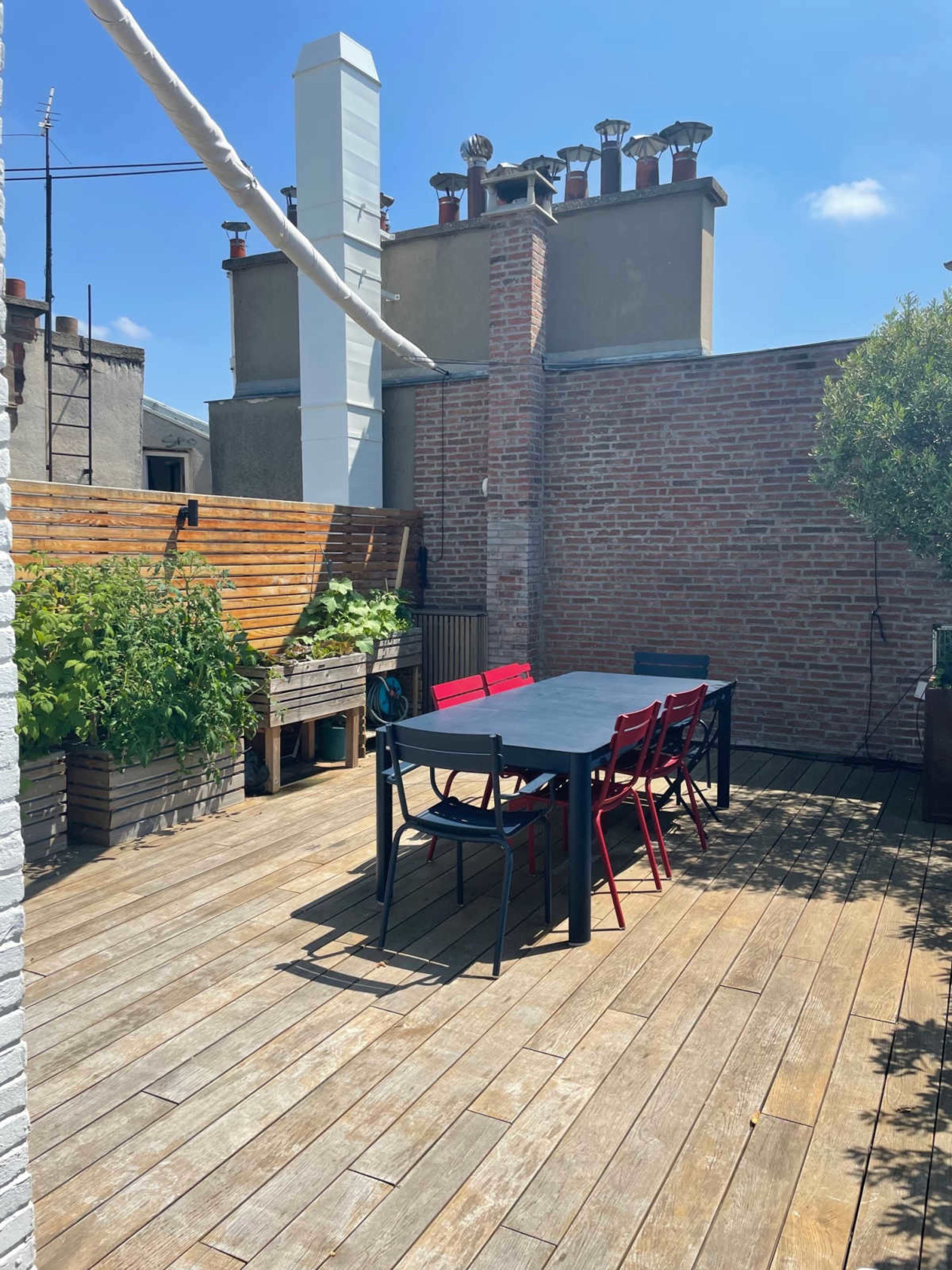 The image shows a wooden rooftop terrace featuring a rectangular black table surrounded by black and red chairs, with greenery in planters against brick walls.