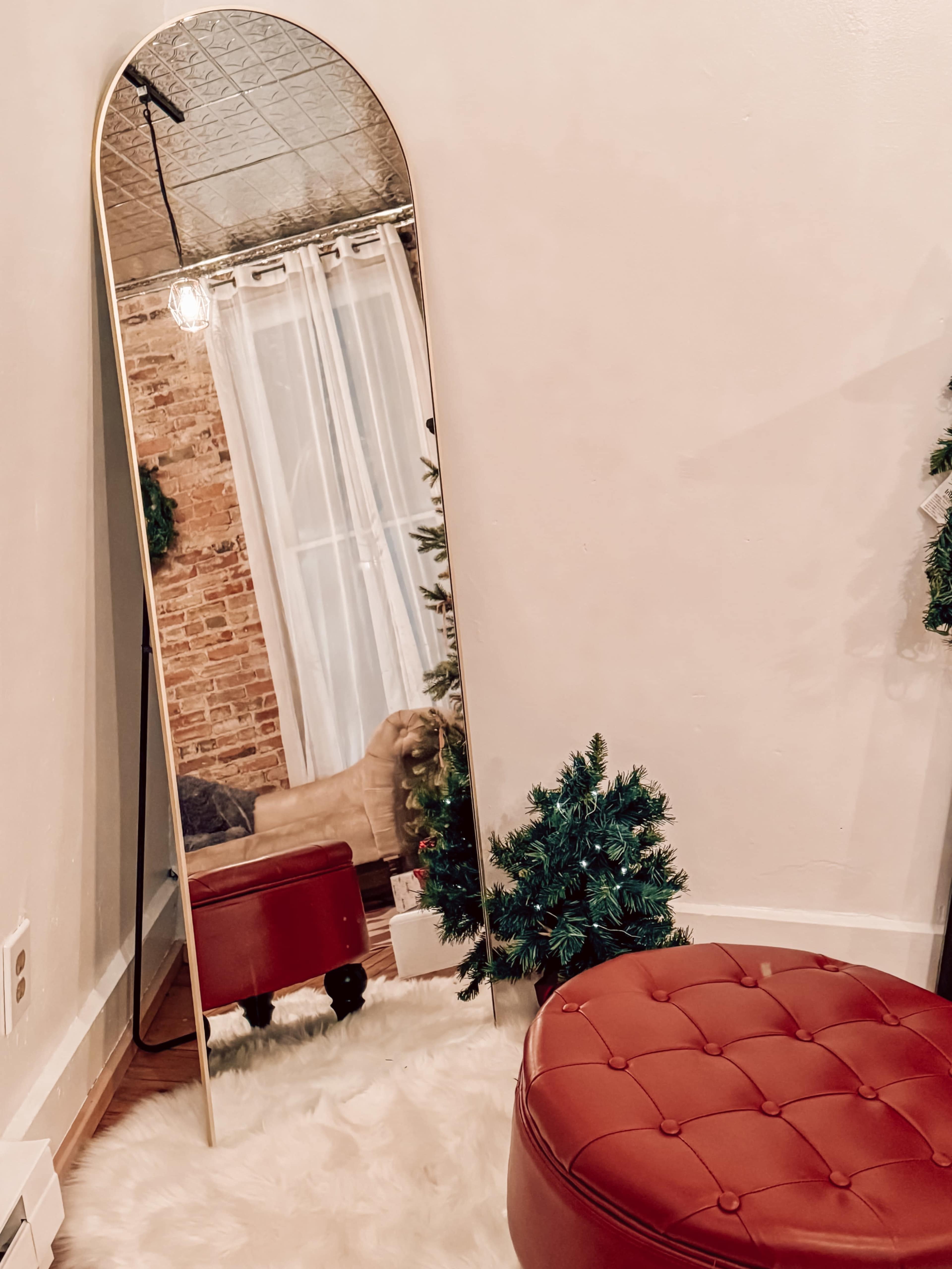 The image shows a tall, arched mirror beside a red upholstered ottoman, with a small green Christmas tree partially visible in the background.