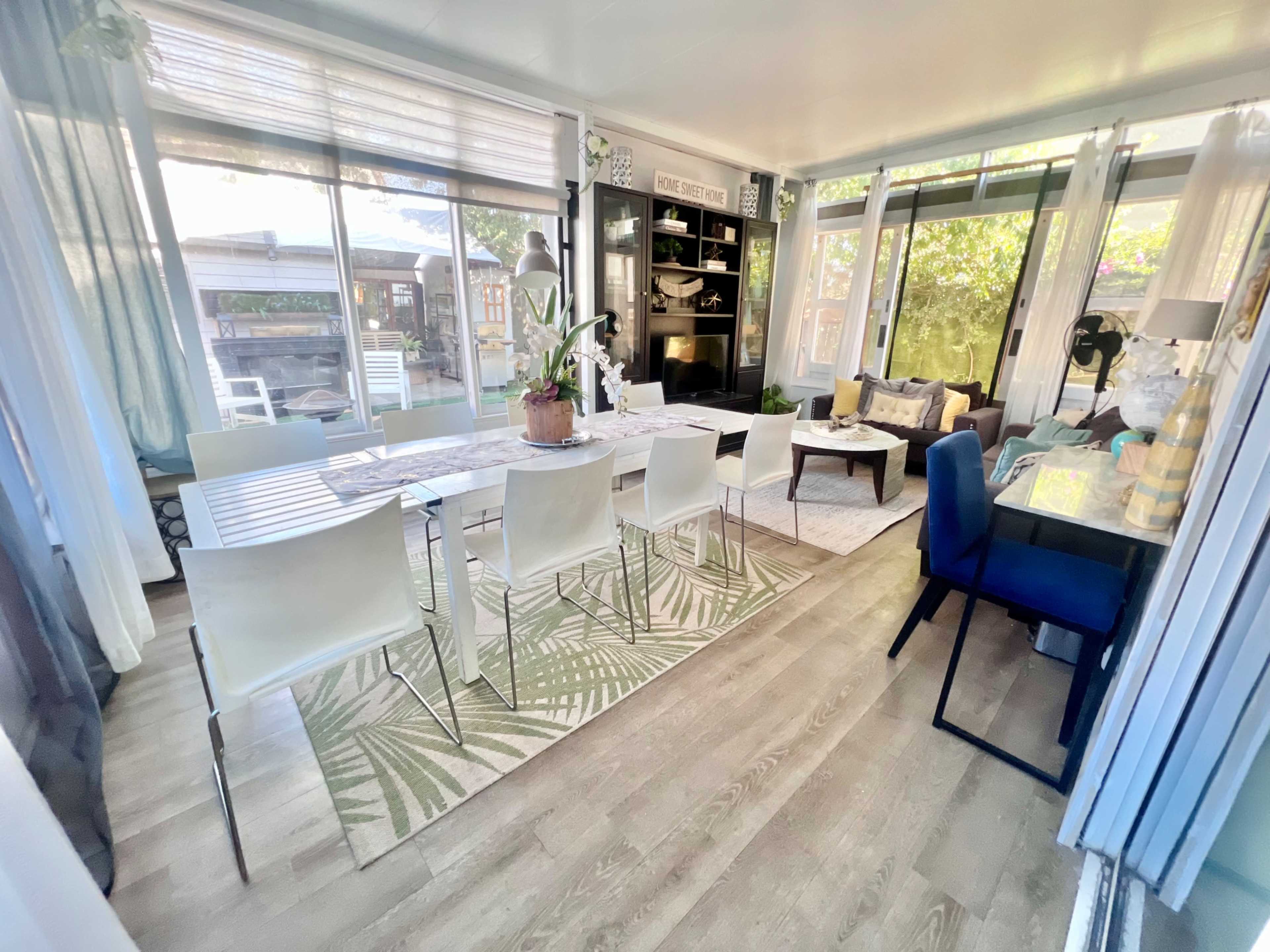 A bright, modern dining area with a long table surrounded by white chairs, and large windows revealing a garden outside.