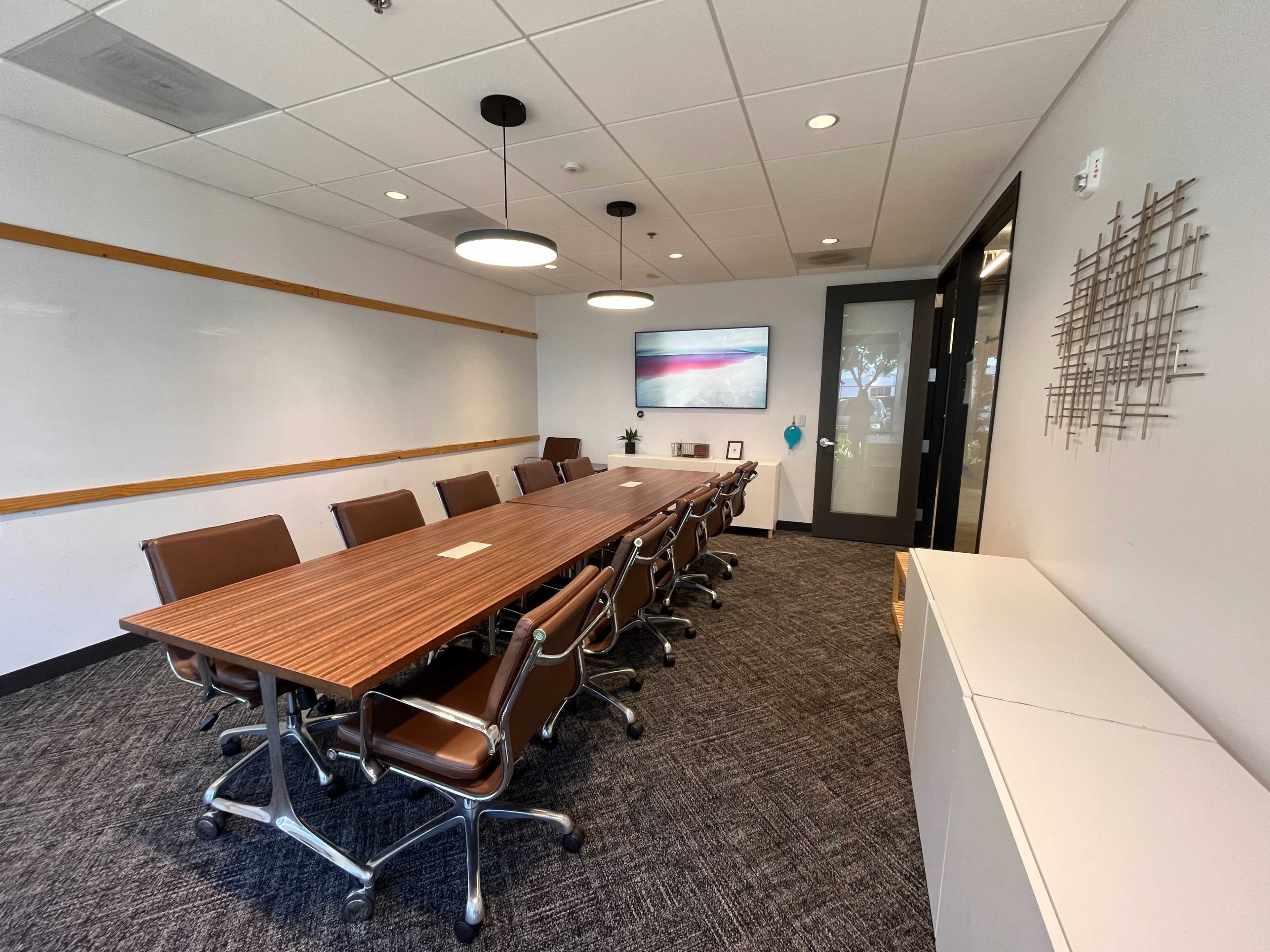 A modern conference room features a long wooden table surrounded by leather chairs, with a whiteboard and a screen on the wall.