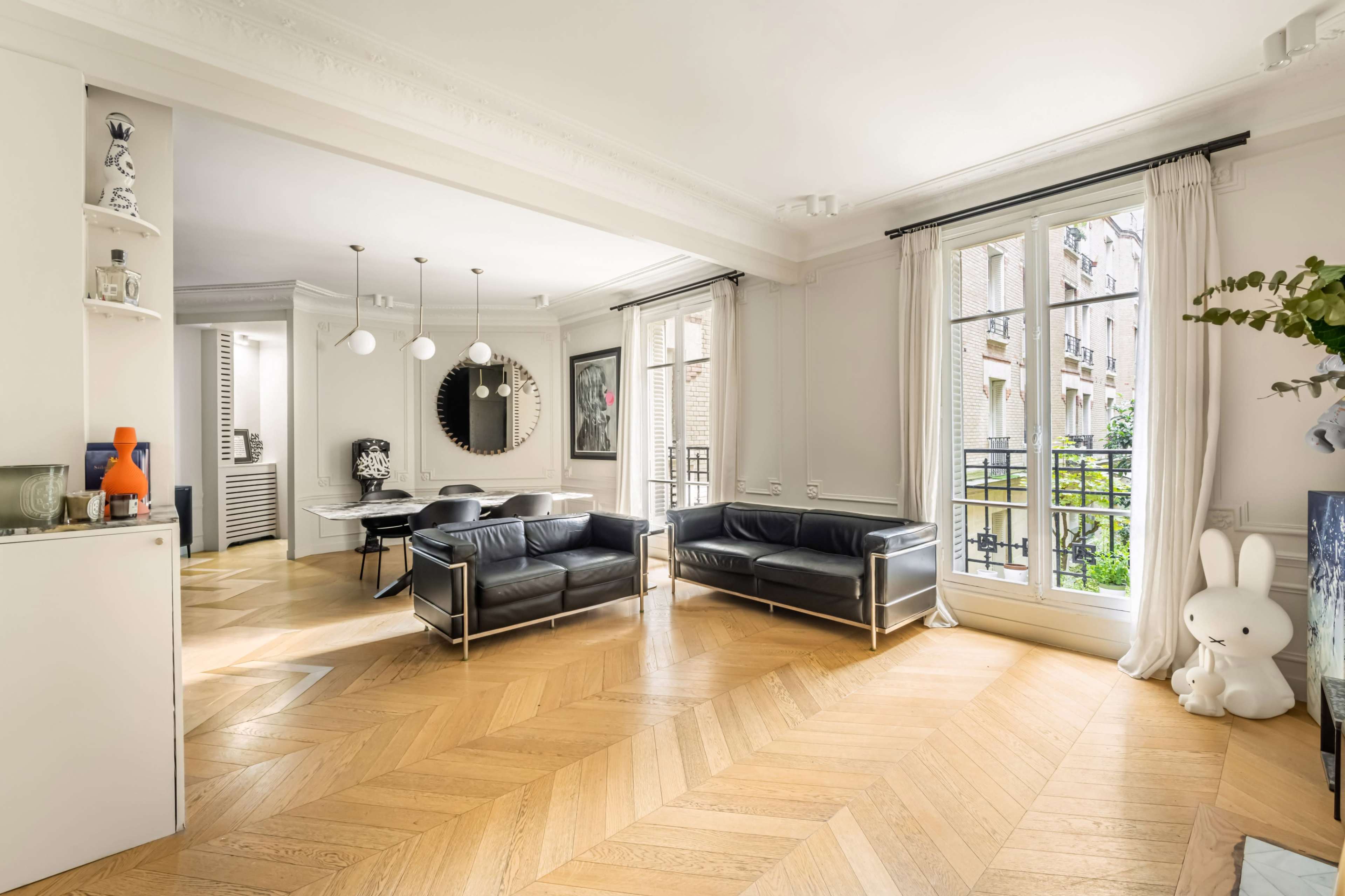 A spacious living room features hardwood flooring, two black sofas, a dining area, and large windows allowing natural light to fill the space.